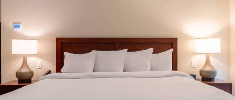 A neatly made bed with a wooden headboard, two bedside lamps, and a wall-mounted thermostat.