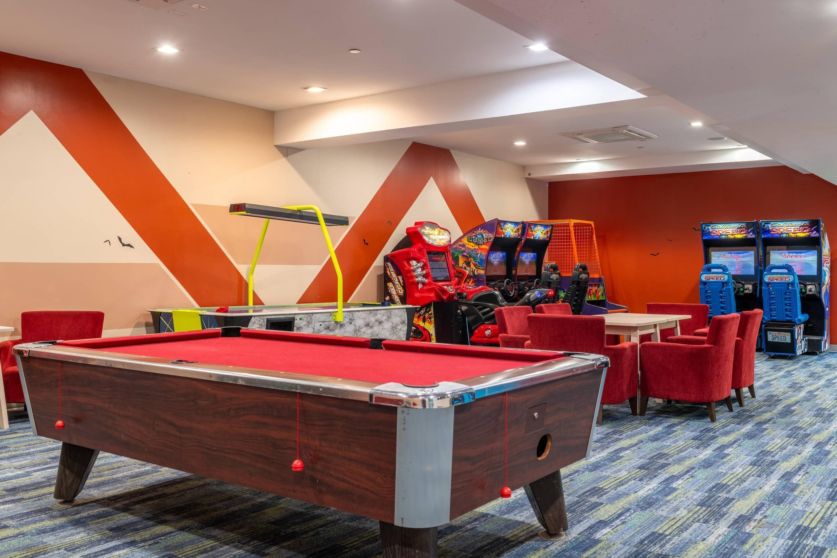 Una mesa de billar con fieltro rojo, sillas rojas y máquinas de arcade en una habitación con paredes naranja y blancas.