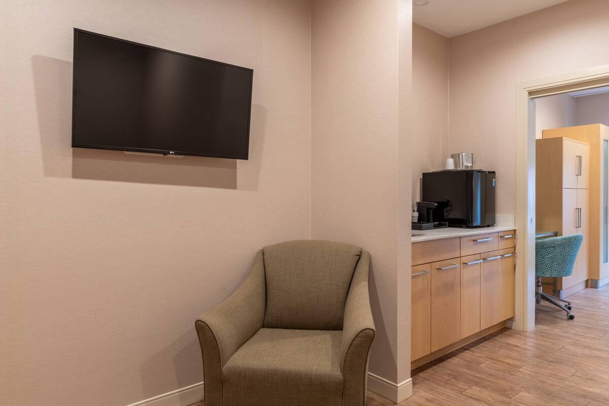 A room with a flat-screen TV mounted on the wall, a beige armchair, a kitchenette with a refrigerator, and a patterned chair.