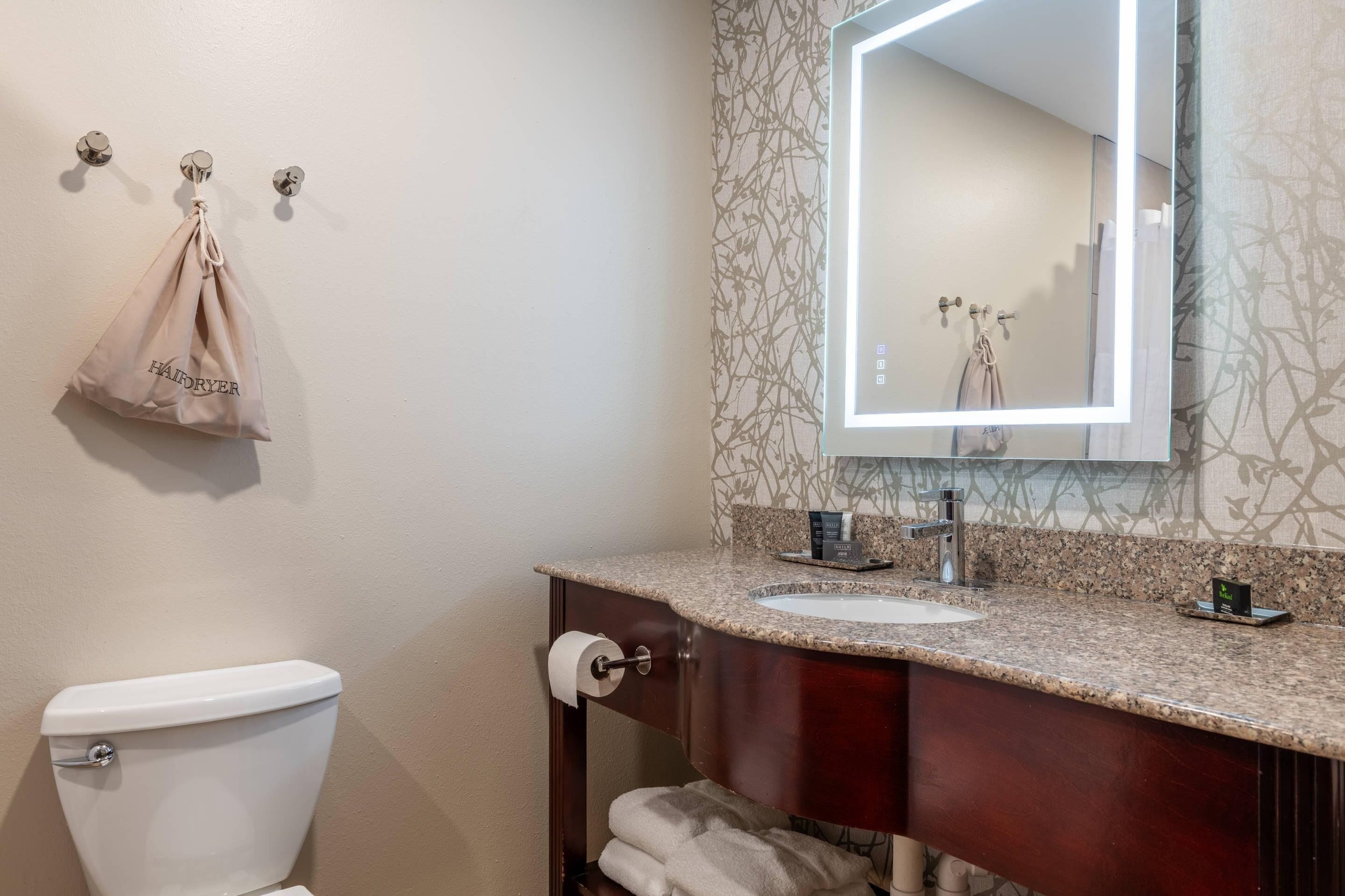 A bathroom with a toilet, sink, mirror, towel rack, and a hanging bag labeled 'HAIRDRYER'.