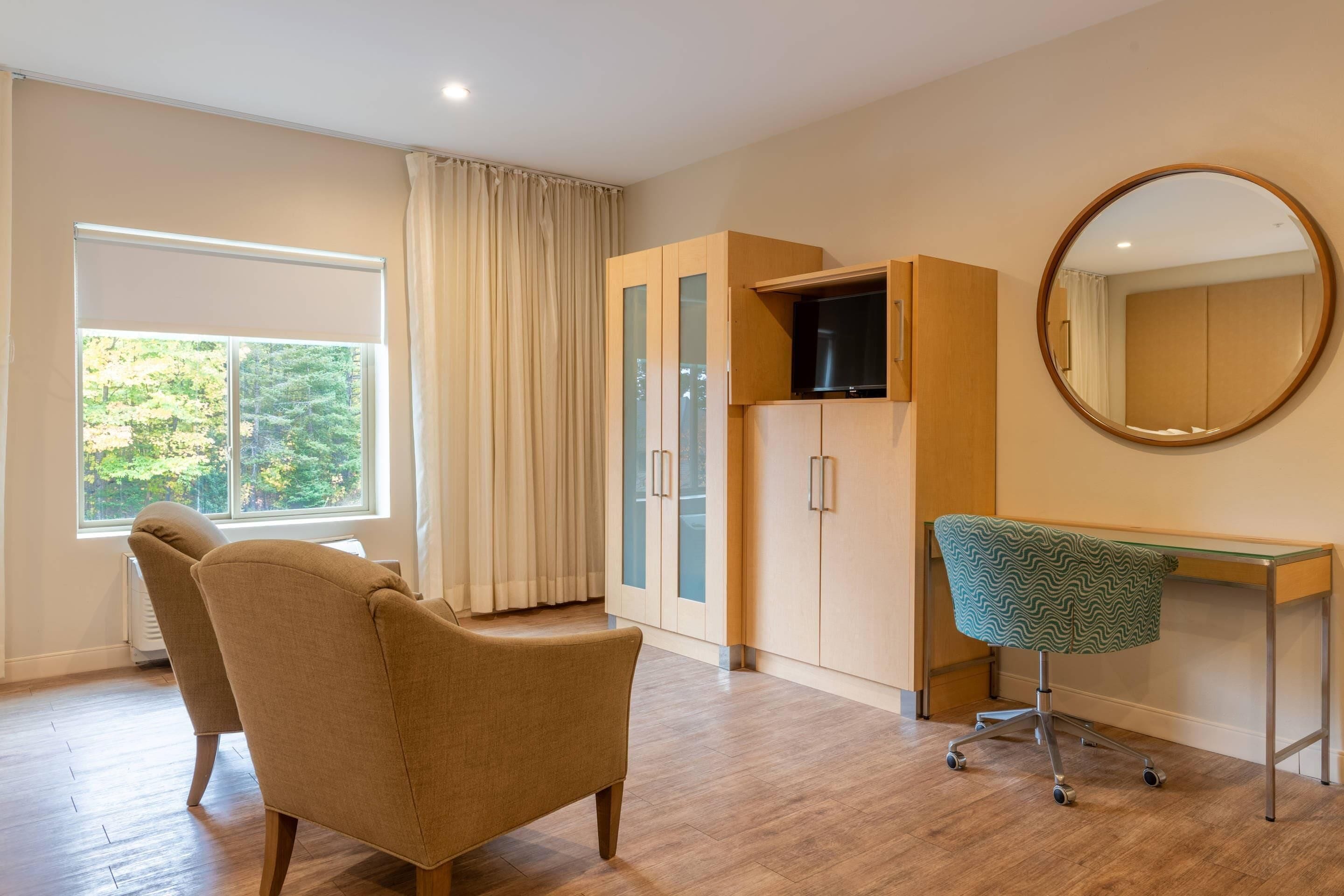 A room with a wooden desk, a green patterned chair, a large mirror, and a wardrobe with glass doors.