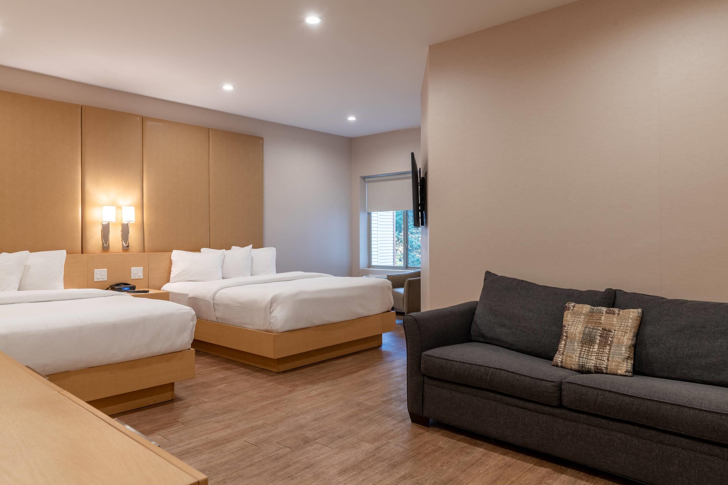 A modern hotel room with two single beds, a sofa, a wooden desk, and a TV mounted on the wall.