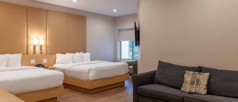A modern hotel room with two single beds, a sofa, a wooden desk, and a TV mounted on the wall.