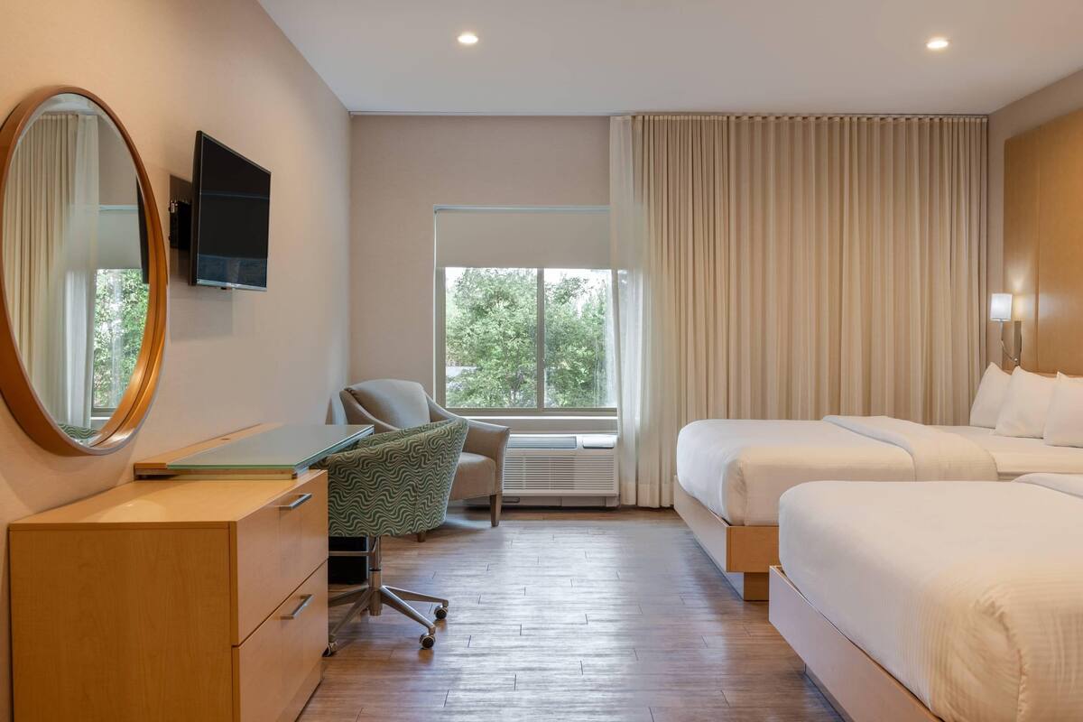 A hotel room with a desk, chair, two beds, a large mirror, and a TV.