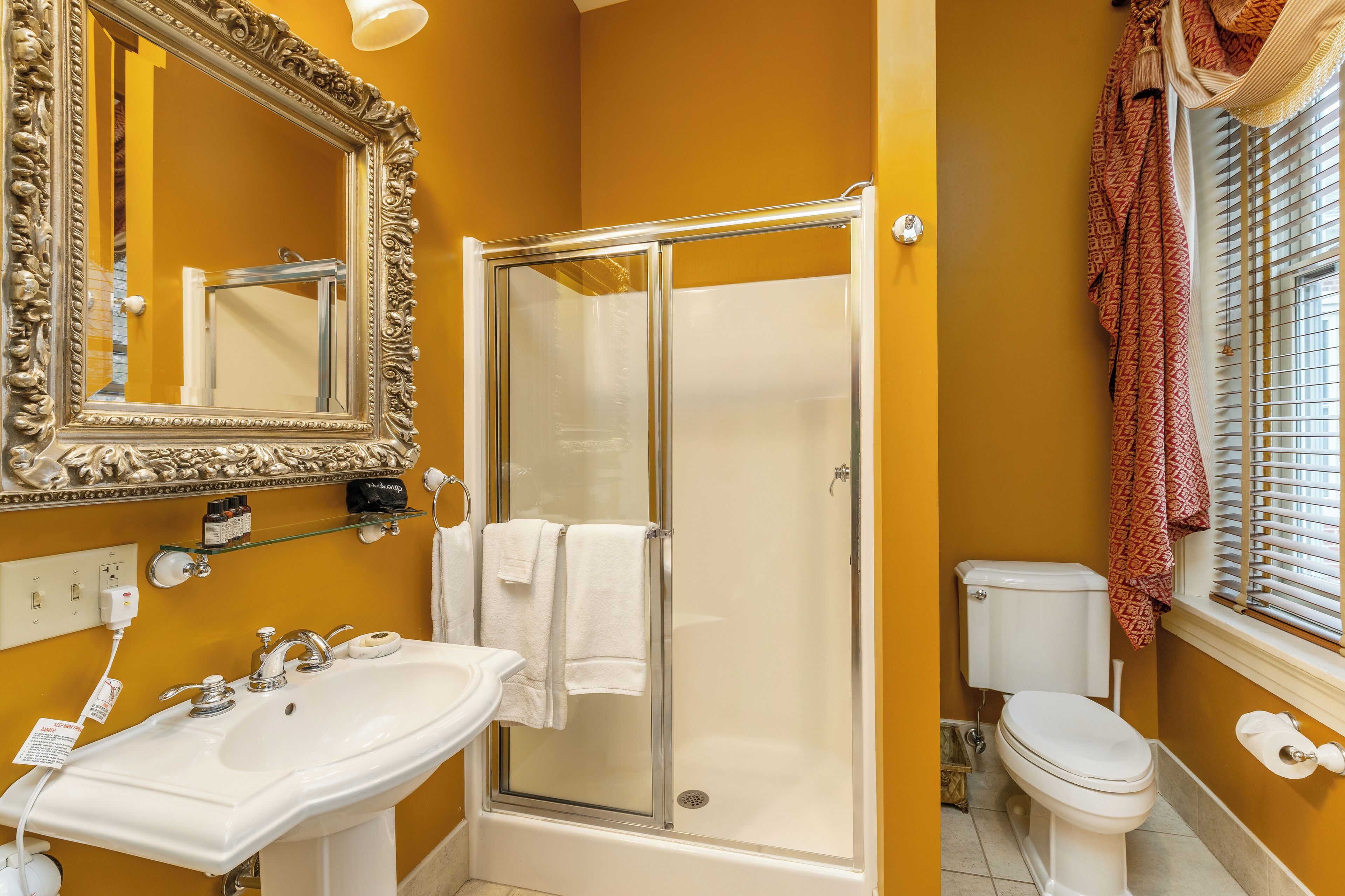 Un baño con ducha envuelta en vidrio, un lavabo blanco, inodoro y un espejo grande y ornamentado.