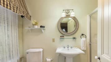 Un baño con inodoro, lavamanos y espejo. Hay una puerta de ducha, un perchero y un estante con artículos de aseo.