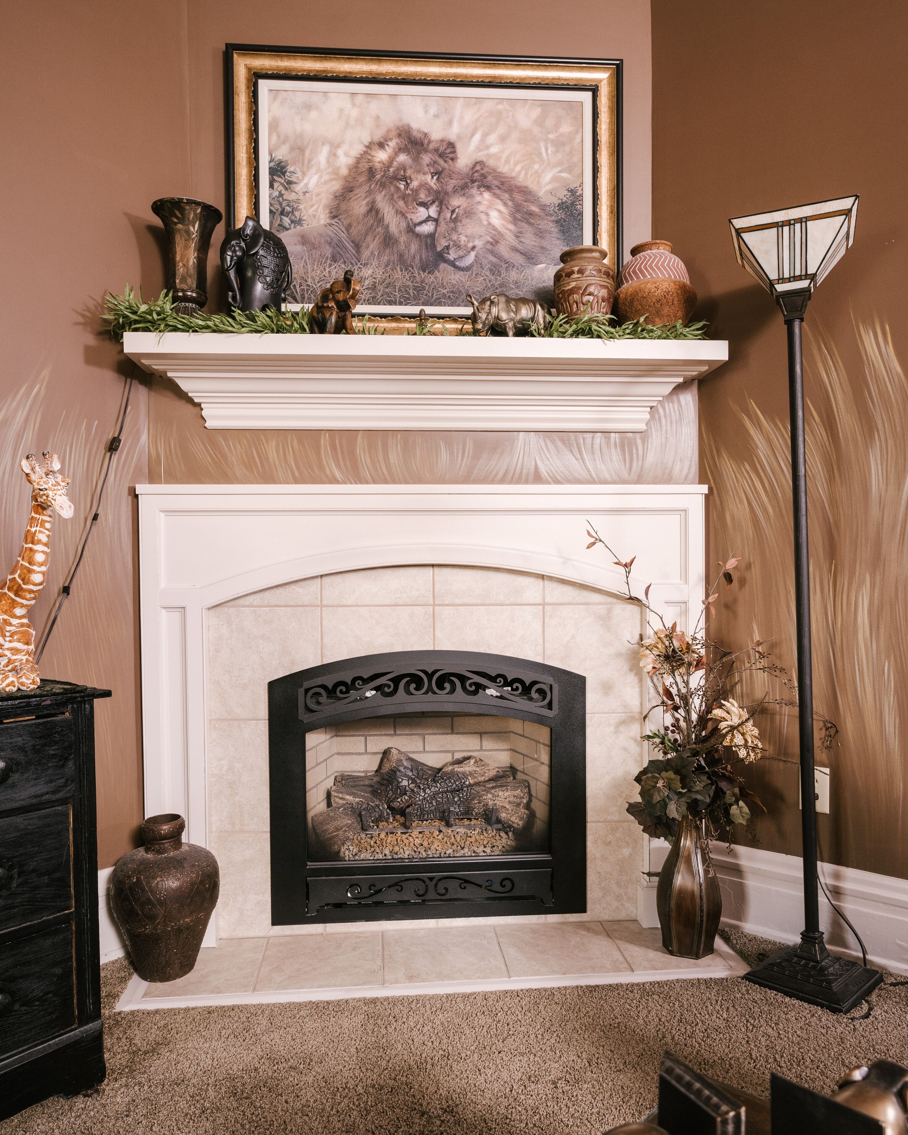 A fireplace with a mantel decorated with various items, a framed picture above, and a vase with dried flowers to the right.