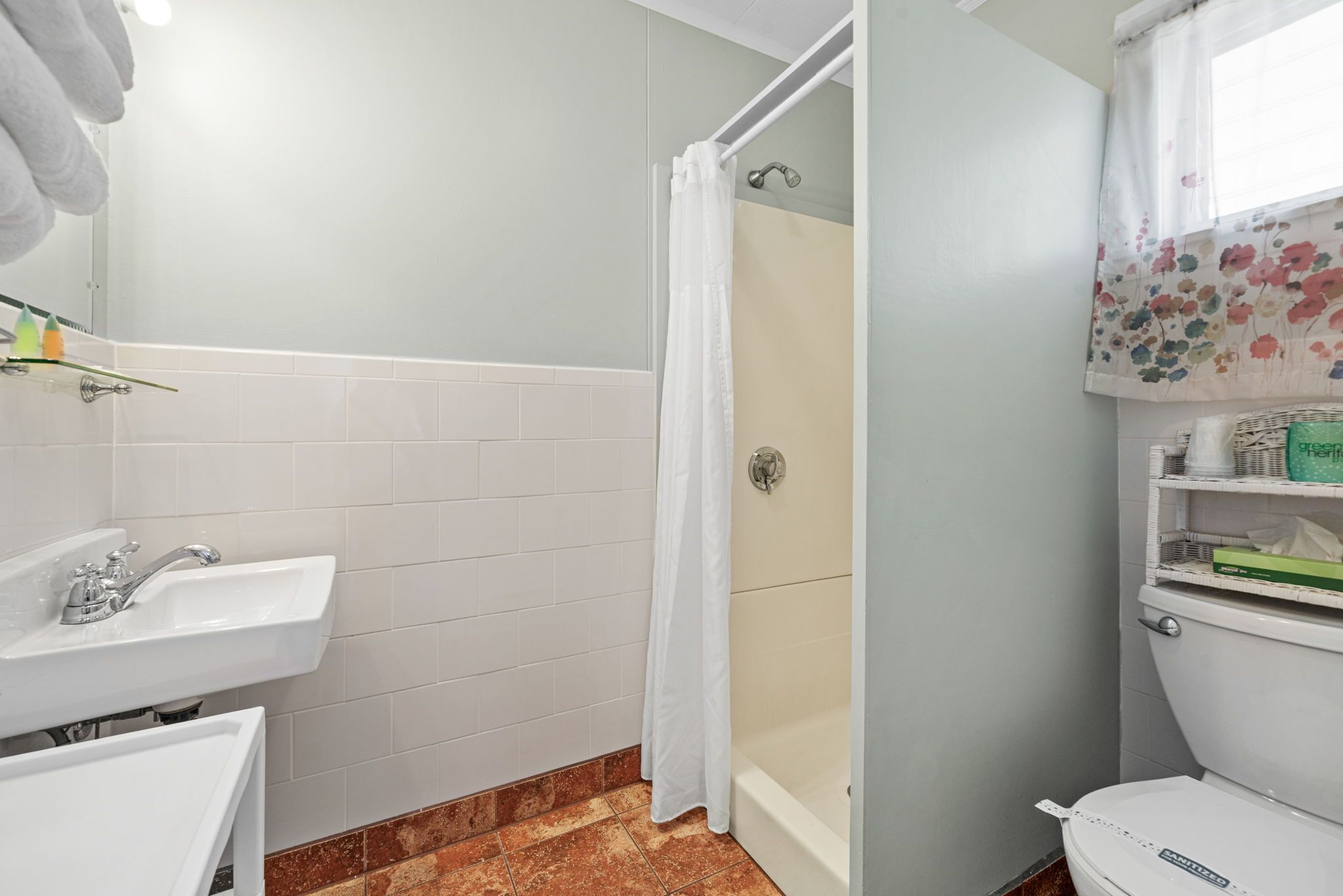 A bathroom with a shower, toilet, sink, and a window with a floral curtain.