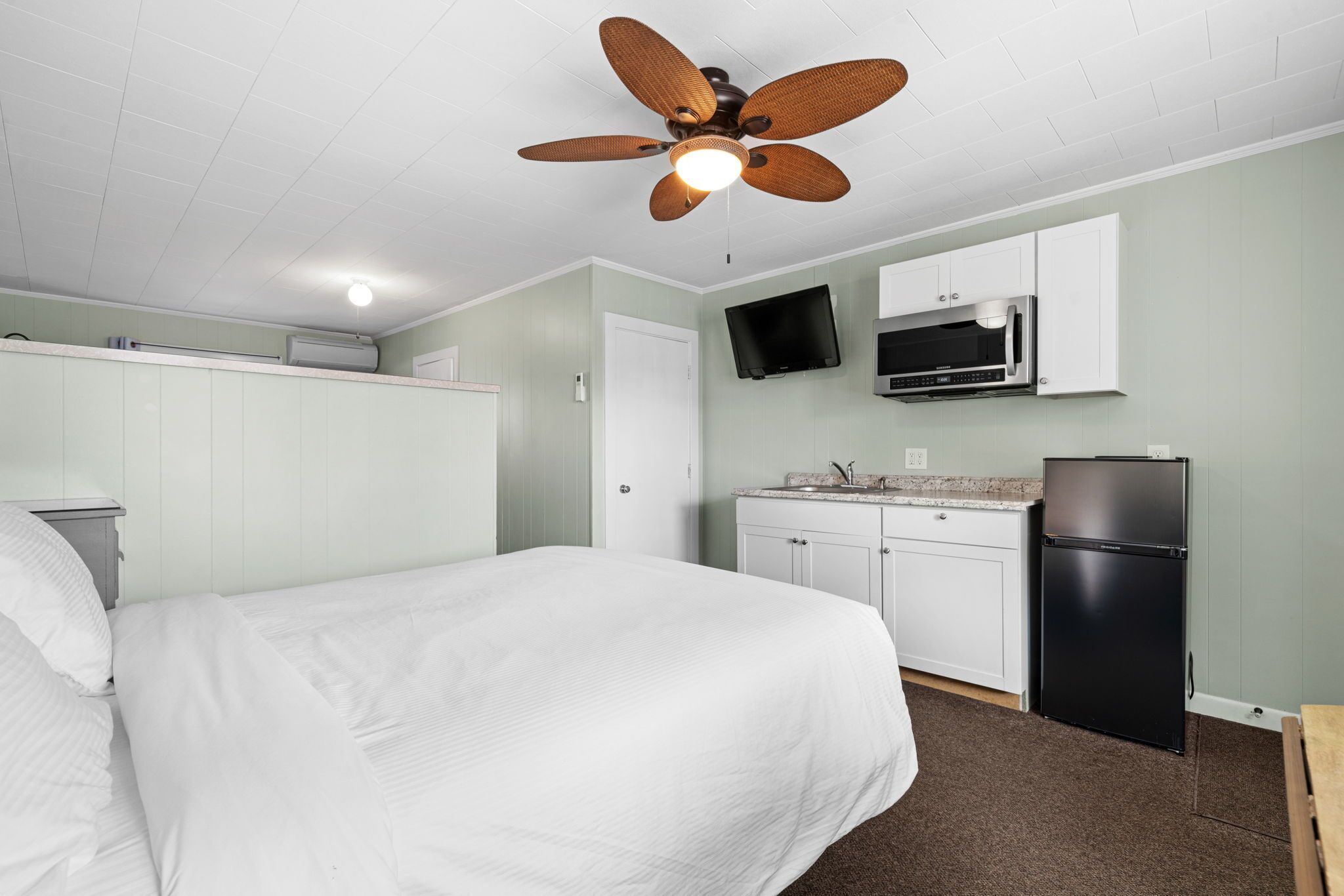 Une chambre de taille réduite comprenant un lit, une kitchenette, un ventilateur de plafond et un téléviseur à écran plat.