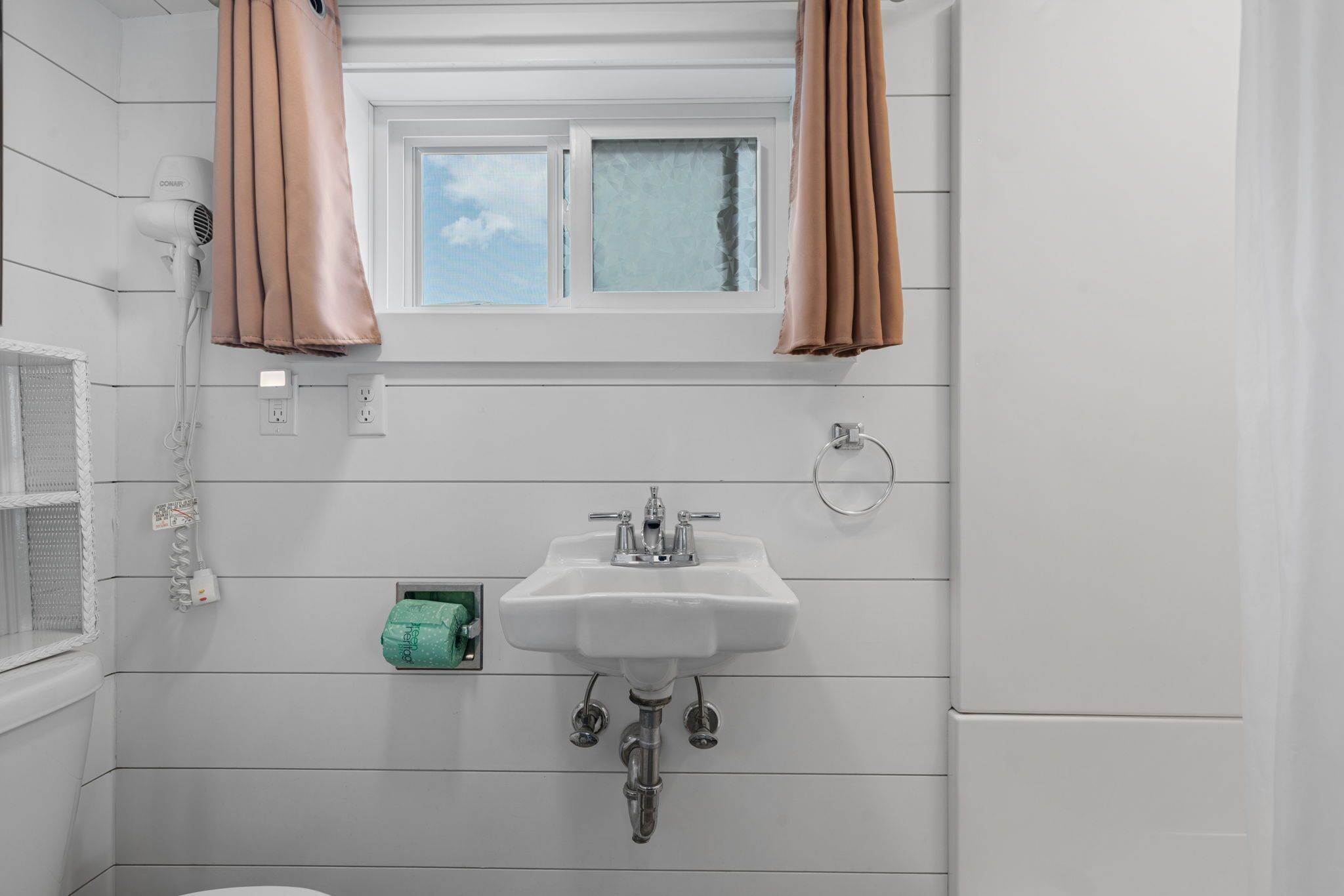 Un baño con lavamanos blanco, secador de pelo y ventana con cortinas.