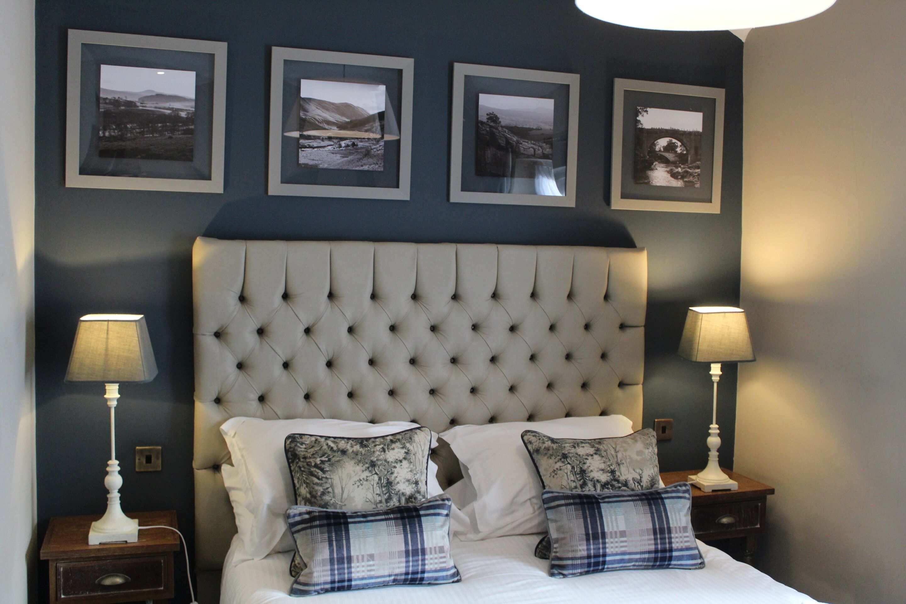 Une chambre à coucher avec une tête de lit capitonnée, deux tables de chevet avec des lampes, un lit avec du linge de lit blanc et des oreillers à carreaux, et quatre photographies encadrées accrochées au mur.