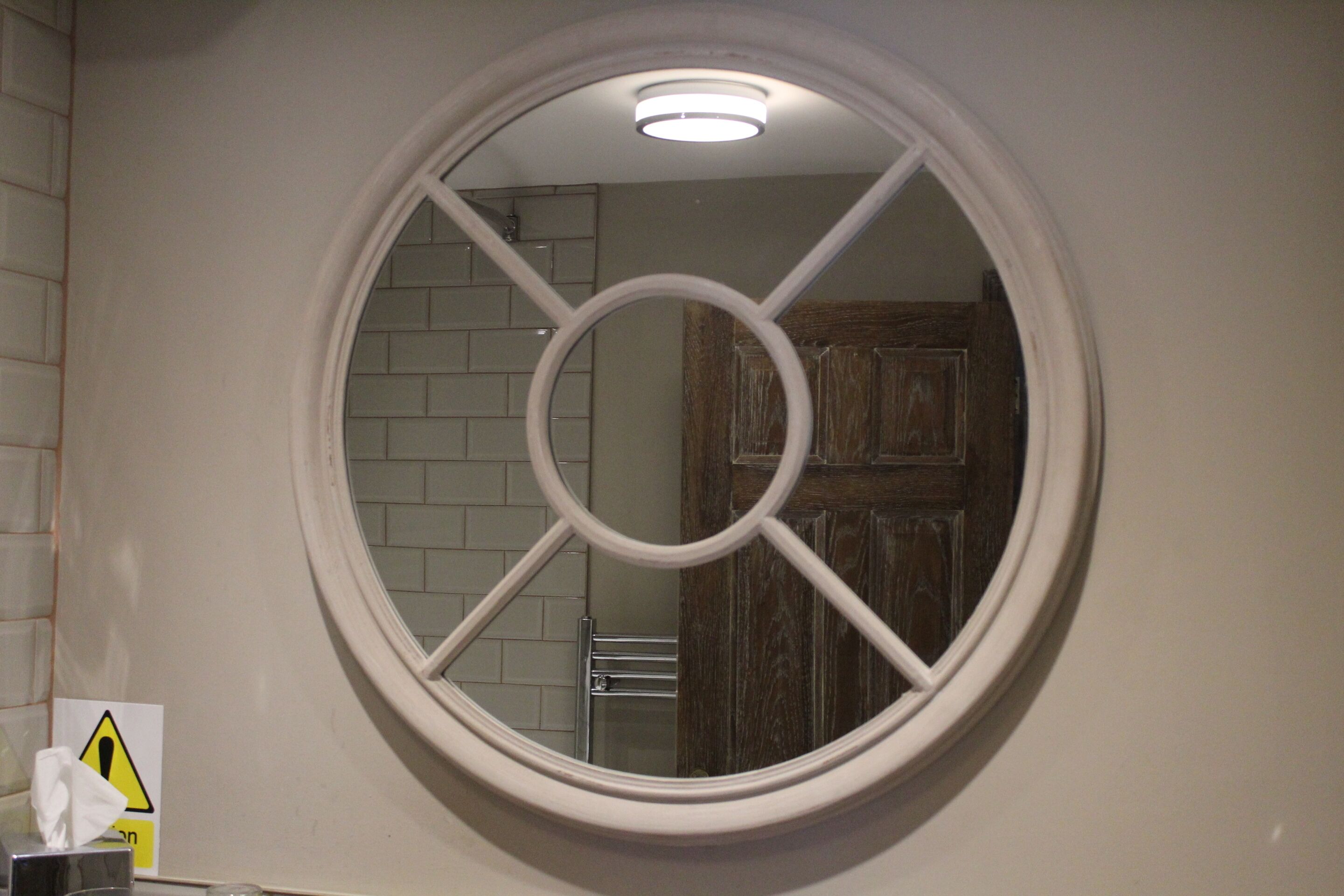 Une salle de bain avec un miroir rond, un distributeur de serviettes et un panneau d’avertissement sur le mur.