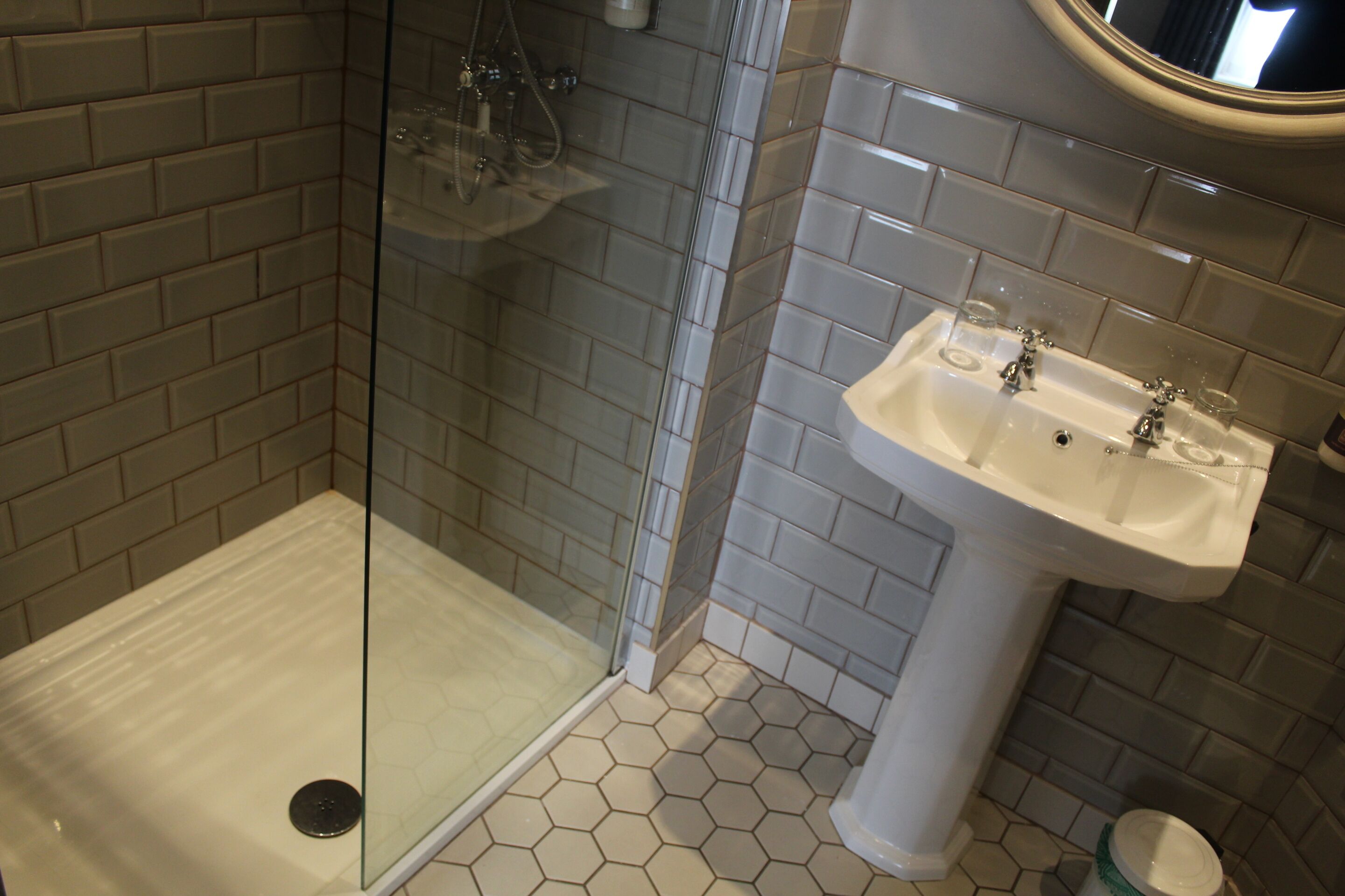 Un baño con ducha acristalada, lavamanos blanco con dos grifos y azulejos de patrón hexagonal.