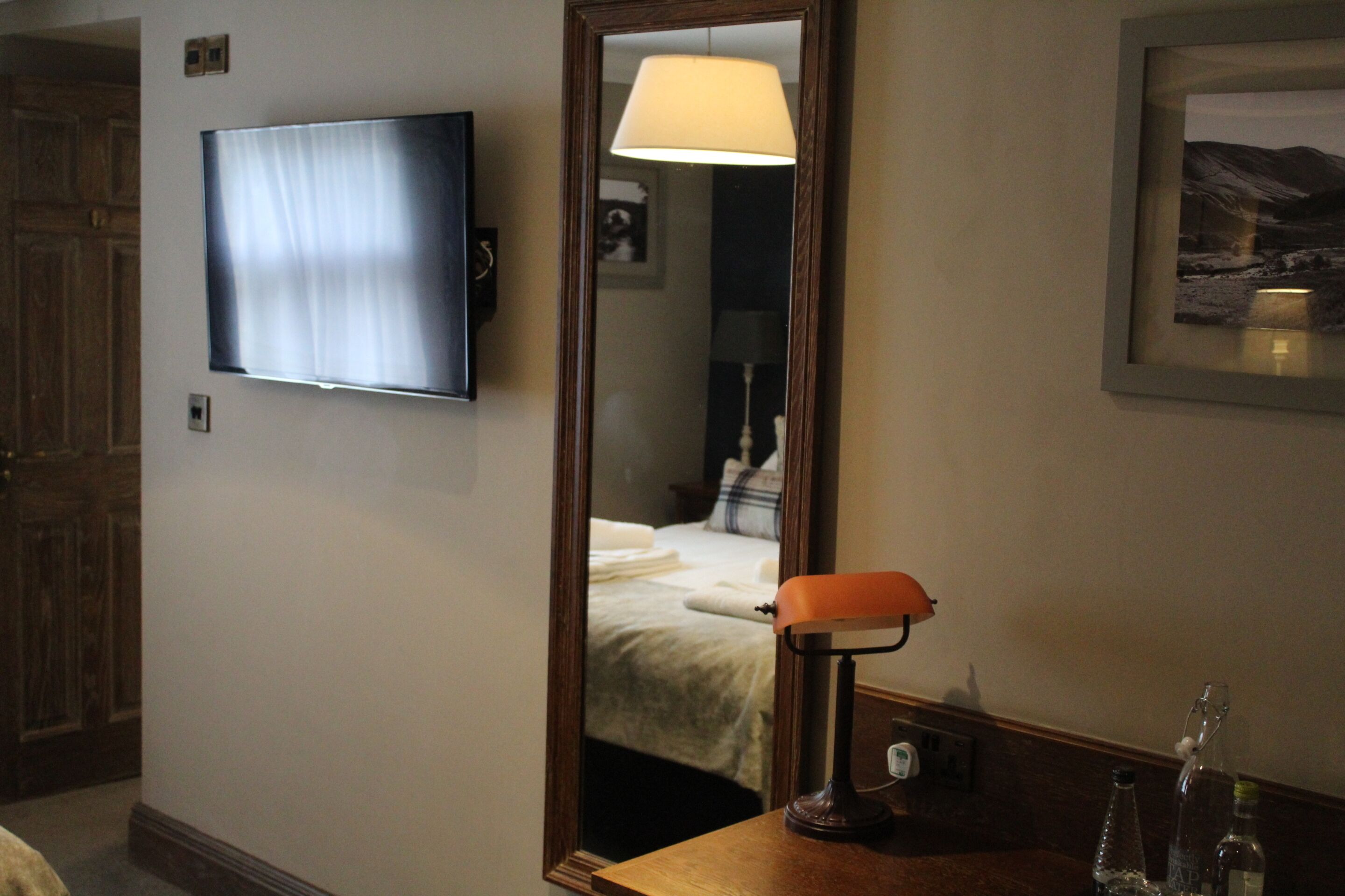 Una camera d'albergo con televisore fissato al muro, una lampada da comodino, un quadro incorniciato e una porta in legno.