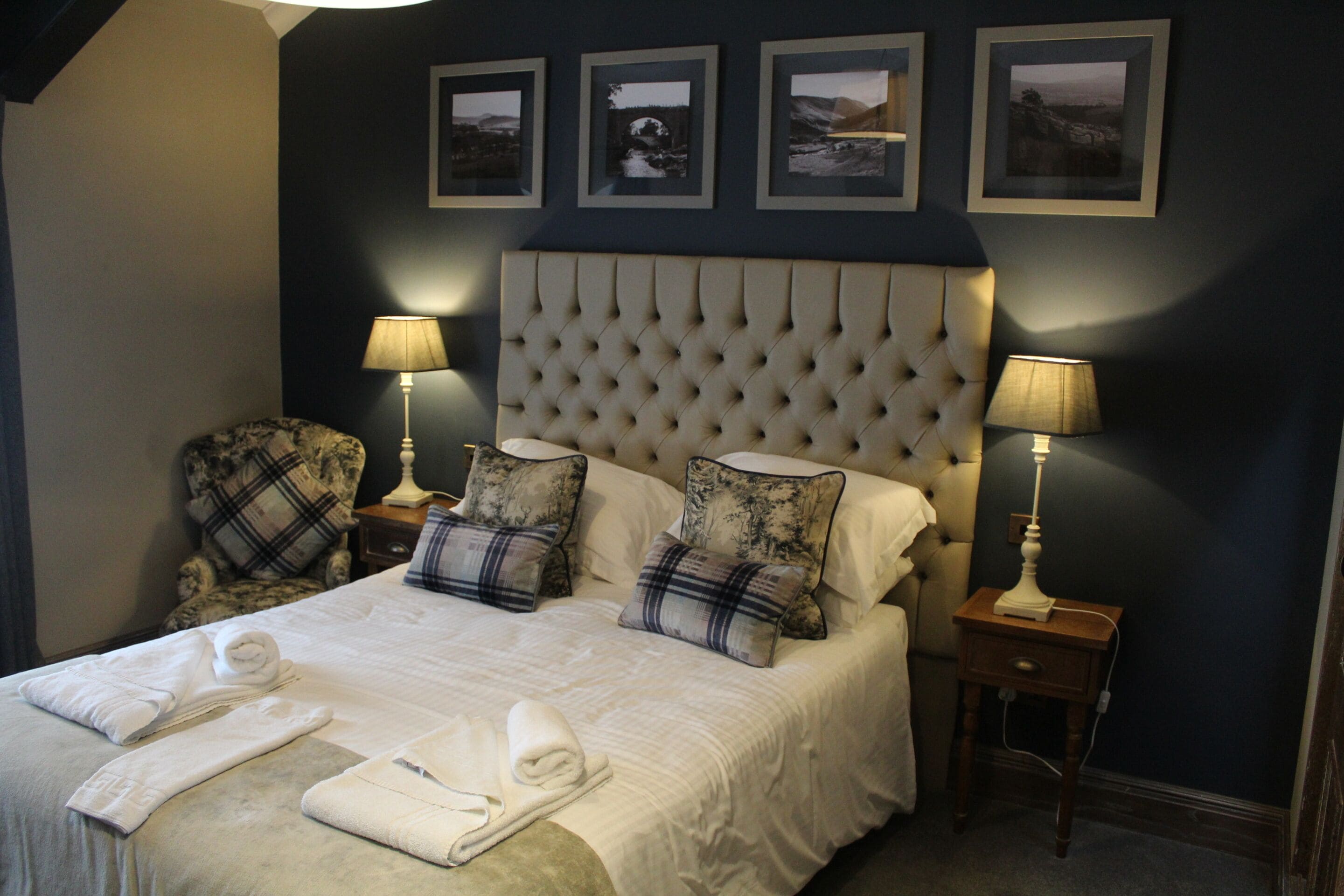 A neatly made bed with white linens and towels, flanked by two nightstands with lamps, in a room with a tufted headboard and framed pictures on the wall.