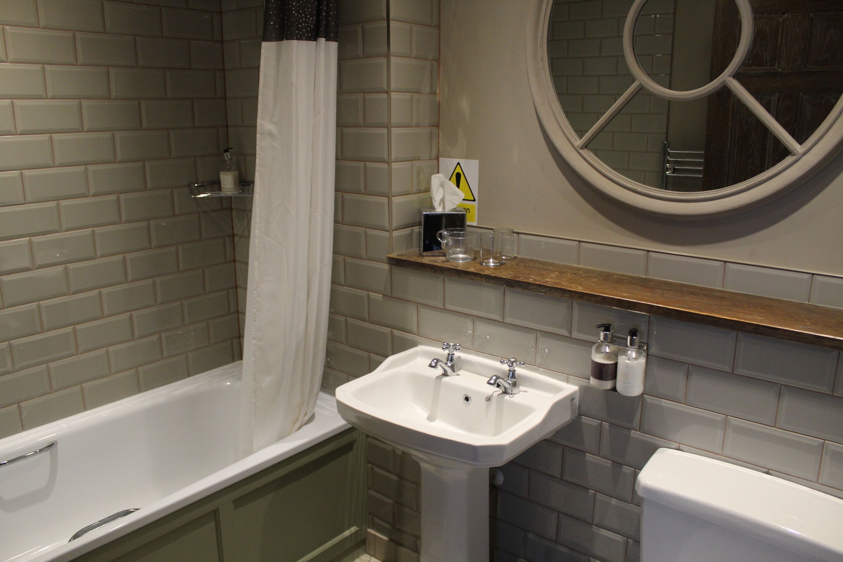 Une salle de bain avec une baignoire, un lavabo et des toilettes. On y trouve une étagère avec des articles de toilette et un panneau d’avertissement.