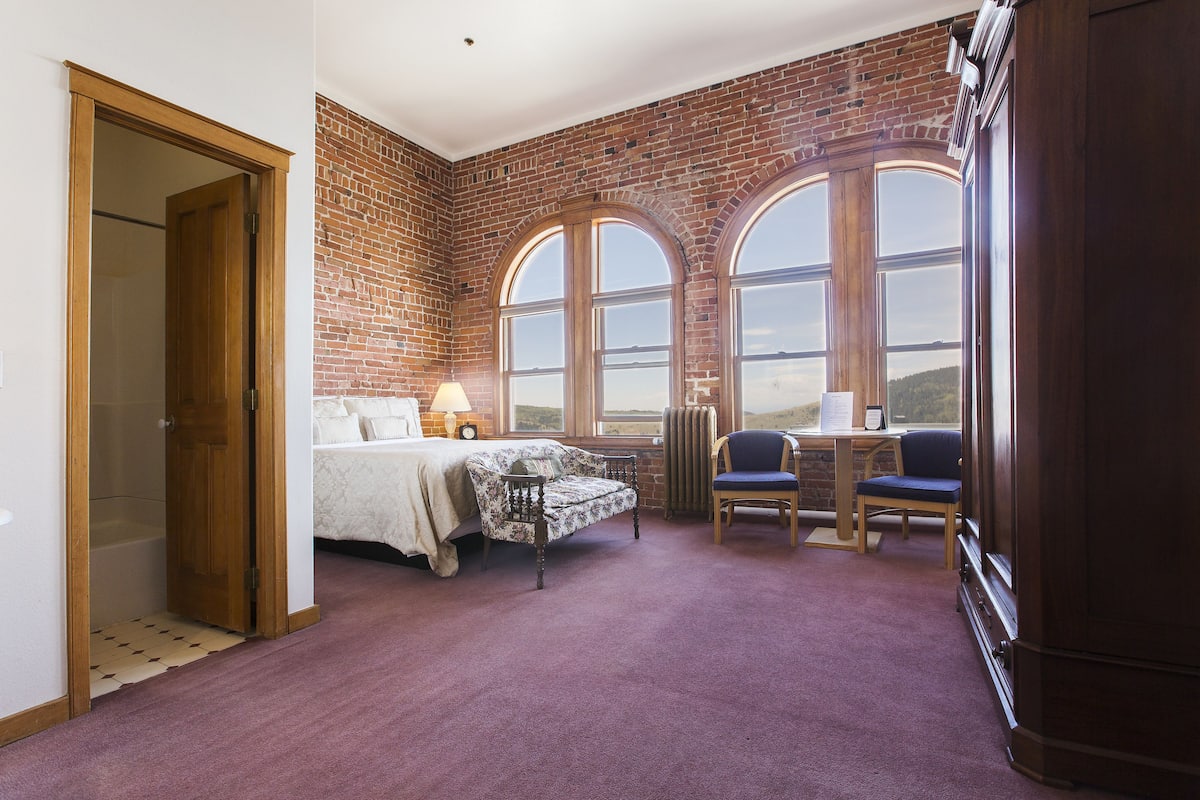 A room with a bed, a desk, and a view through large arched windows.
