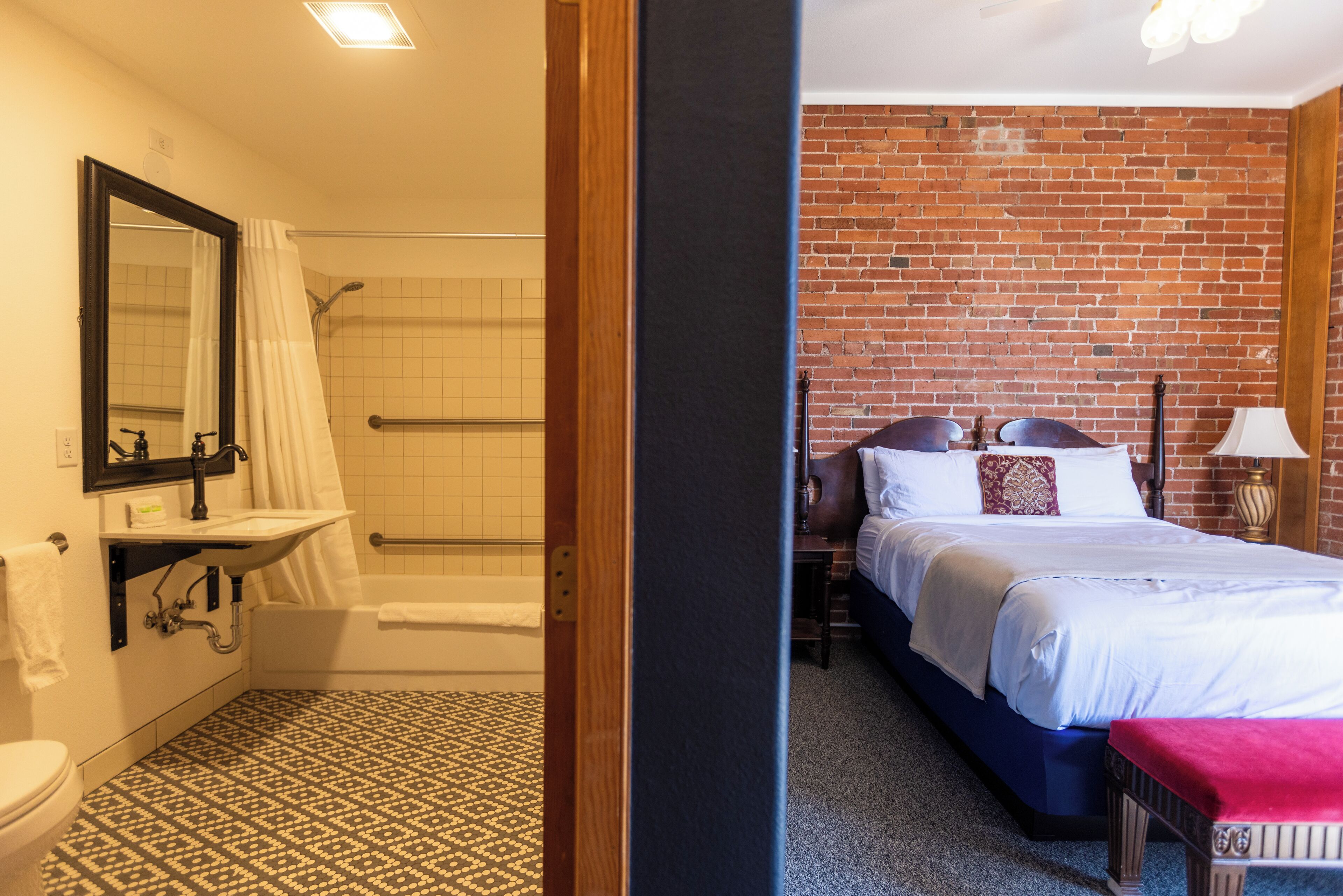 A hotel room with a bathroom featuring a bathtub, a toilet, and a sink. The room has a brick wall and a bed with a red pillow.