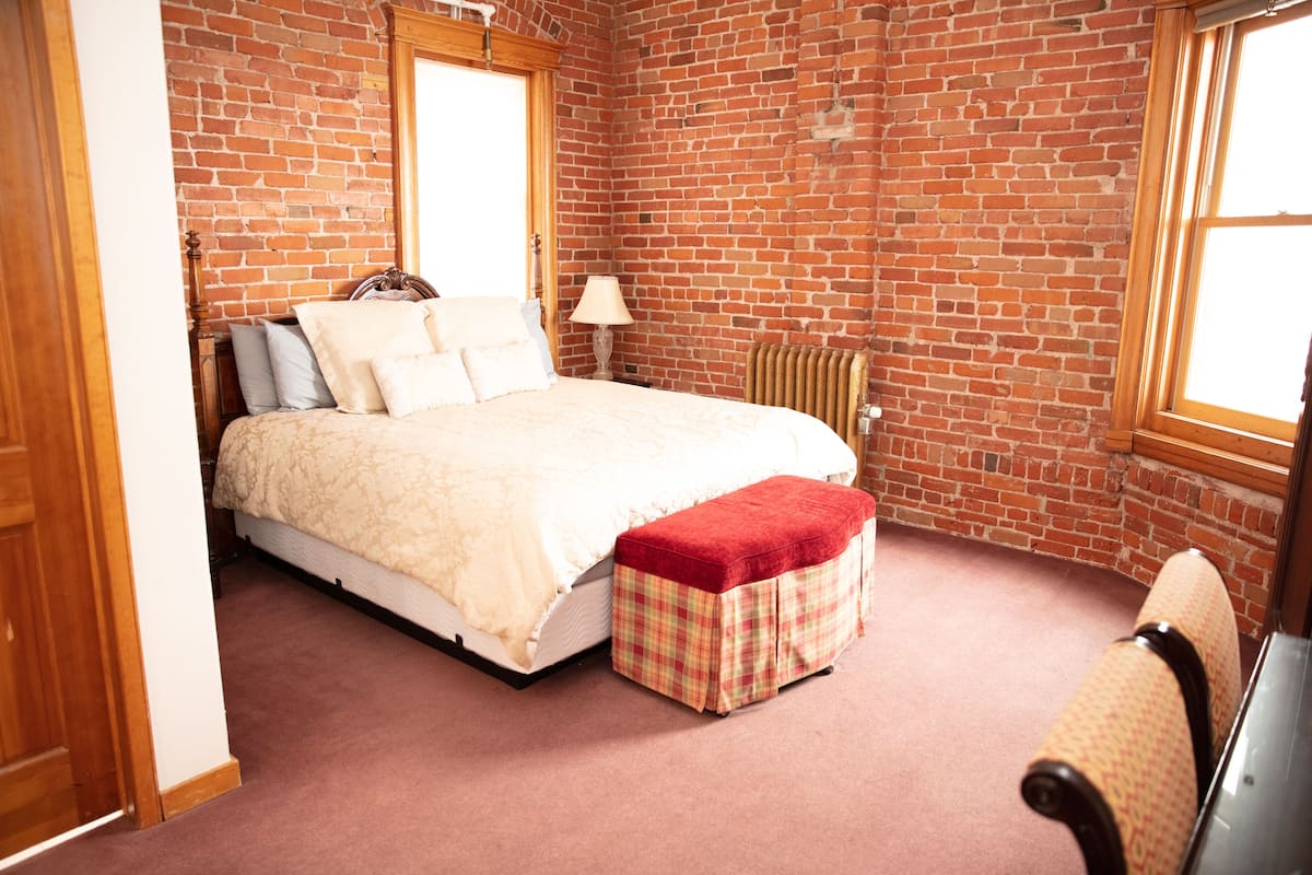 A bedroom with a brick wall, a bed with a red and white checkered ottoman, a wooden headboard, and a radiator.