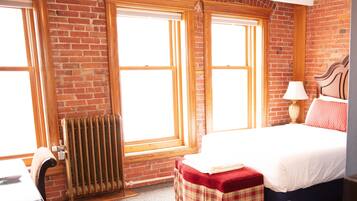 A bedroom with a brick wall, two windows, a radiator, a bed with a red and white pillow, a bedside table, and a lamp.