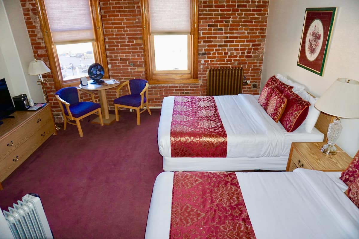 A hotel room with two beds, a desk with a chair, a radiator, and a window with a view of the outside.