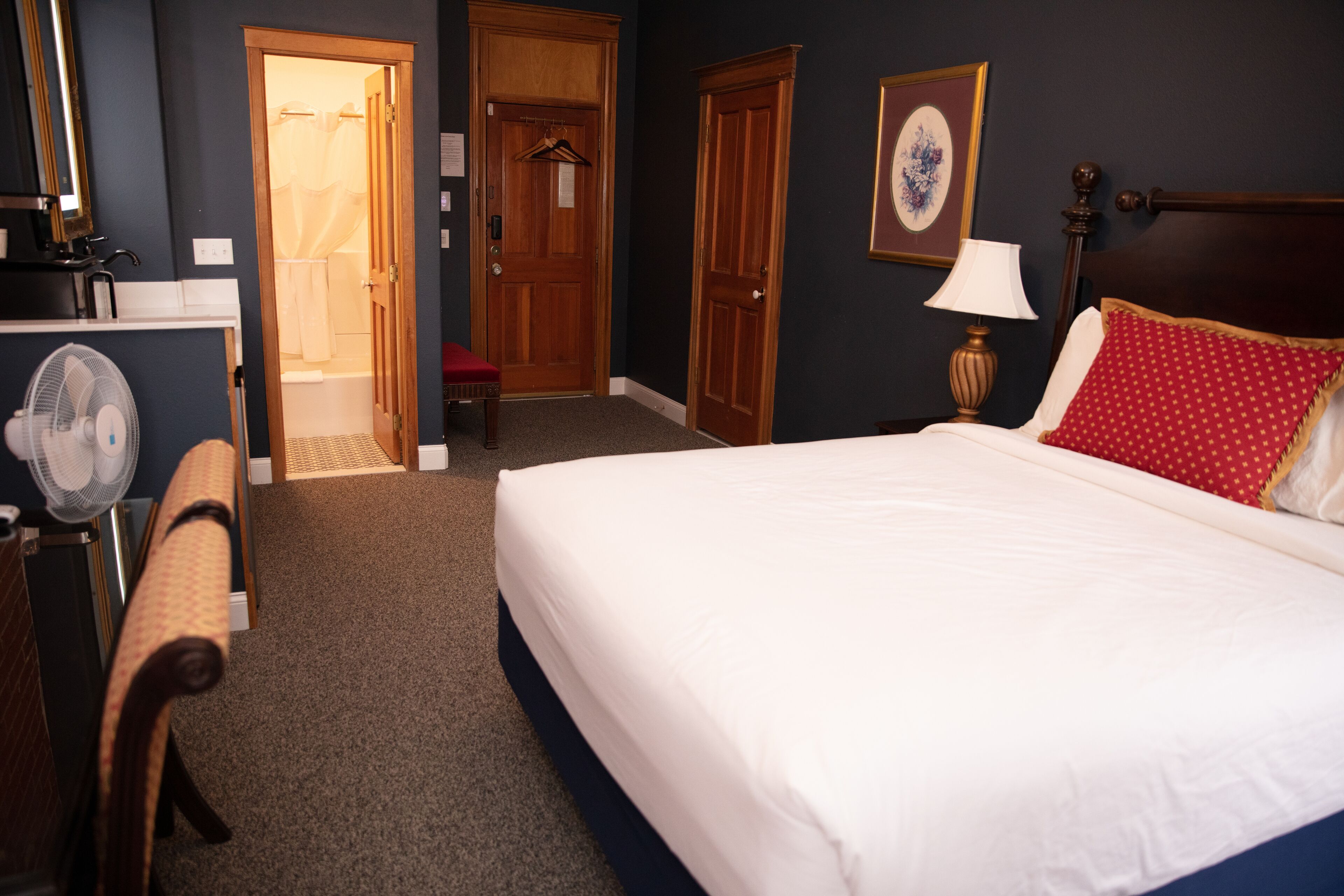 Quarto de hotel com uma cama grande, cabeceira de madeira, mesa de cabeceira com abajur, uma cadeira, escrivaninha com ventilador e banheiro visível por uma porta aberta.