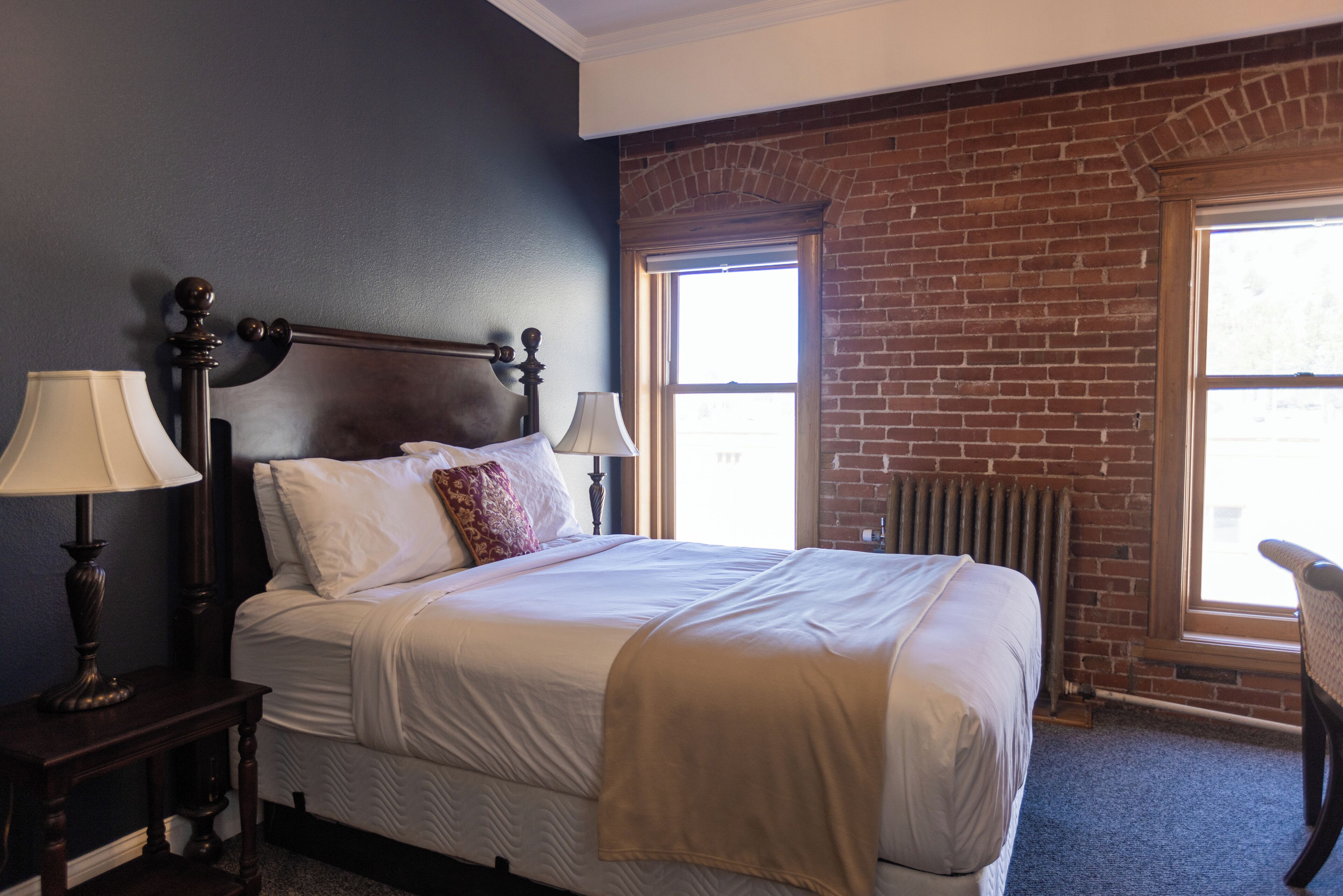 Una cama bien hecha con sábanas blancas y un edredón beige, flanqueada por dos lámparas de noche. Se ve una pared de ladrillo y una ventana con persianas en el fondo.