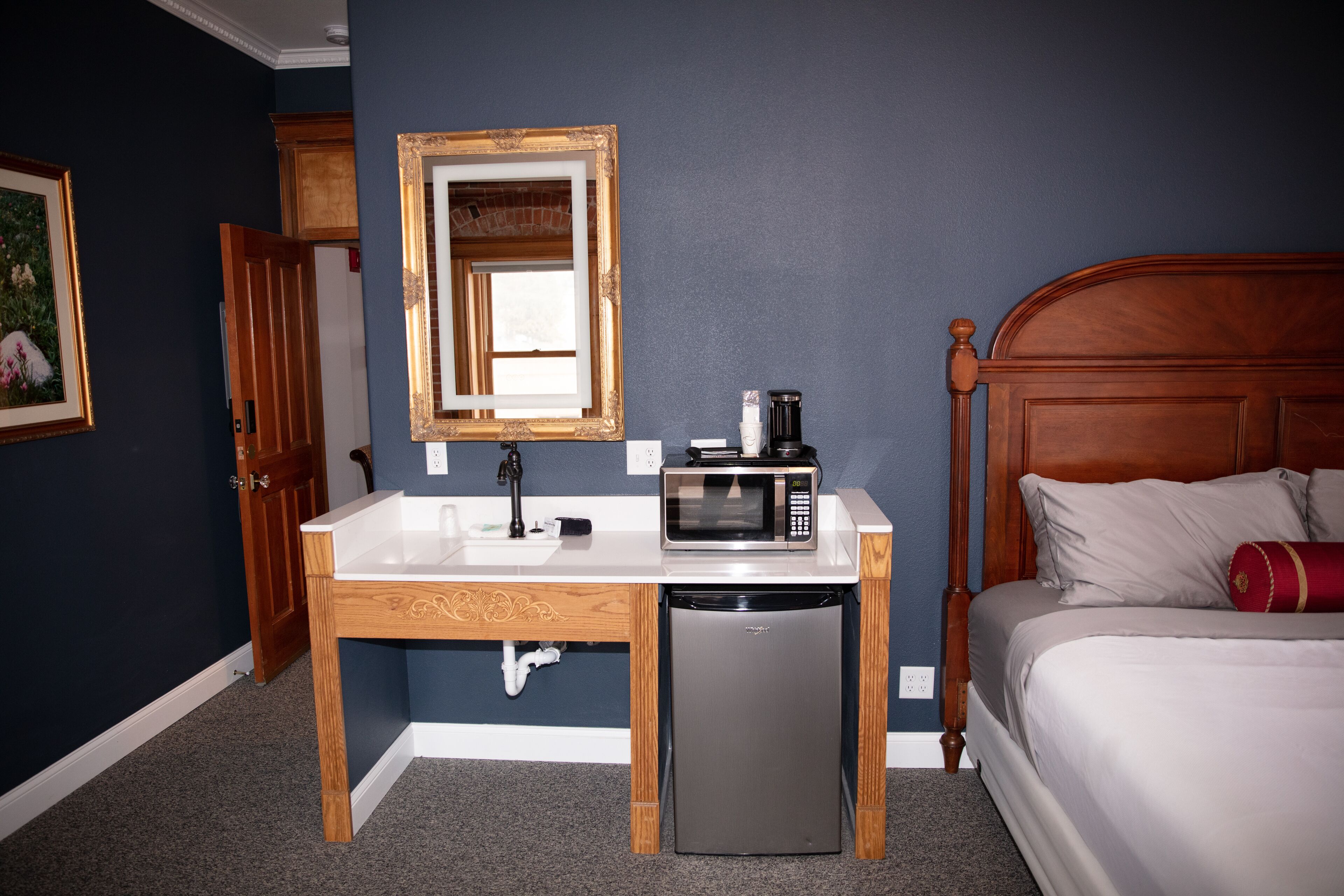 Een compacte hotelkamer met een houten bureau, een magnetron, een kleine koelkast, een bed met houten hoofdeinde en een spiegel aan de muur.
