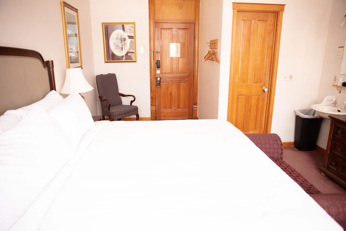A hotel room with a large bed, a chair, a wooden door, and a small bathroom with a sink and mirror.