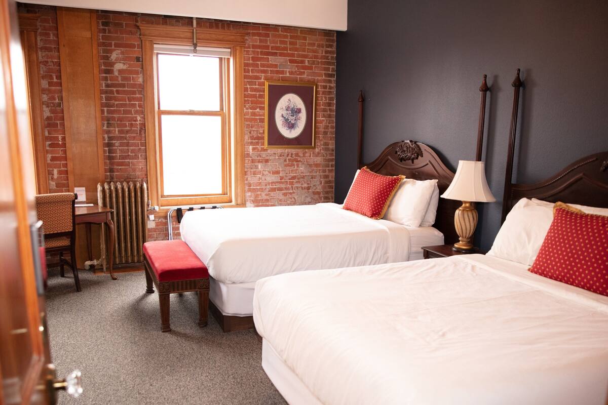 A hotel room with two beds, a wooden chair, a table, and a radiator.