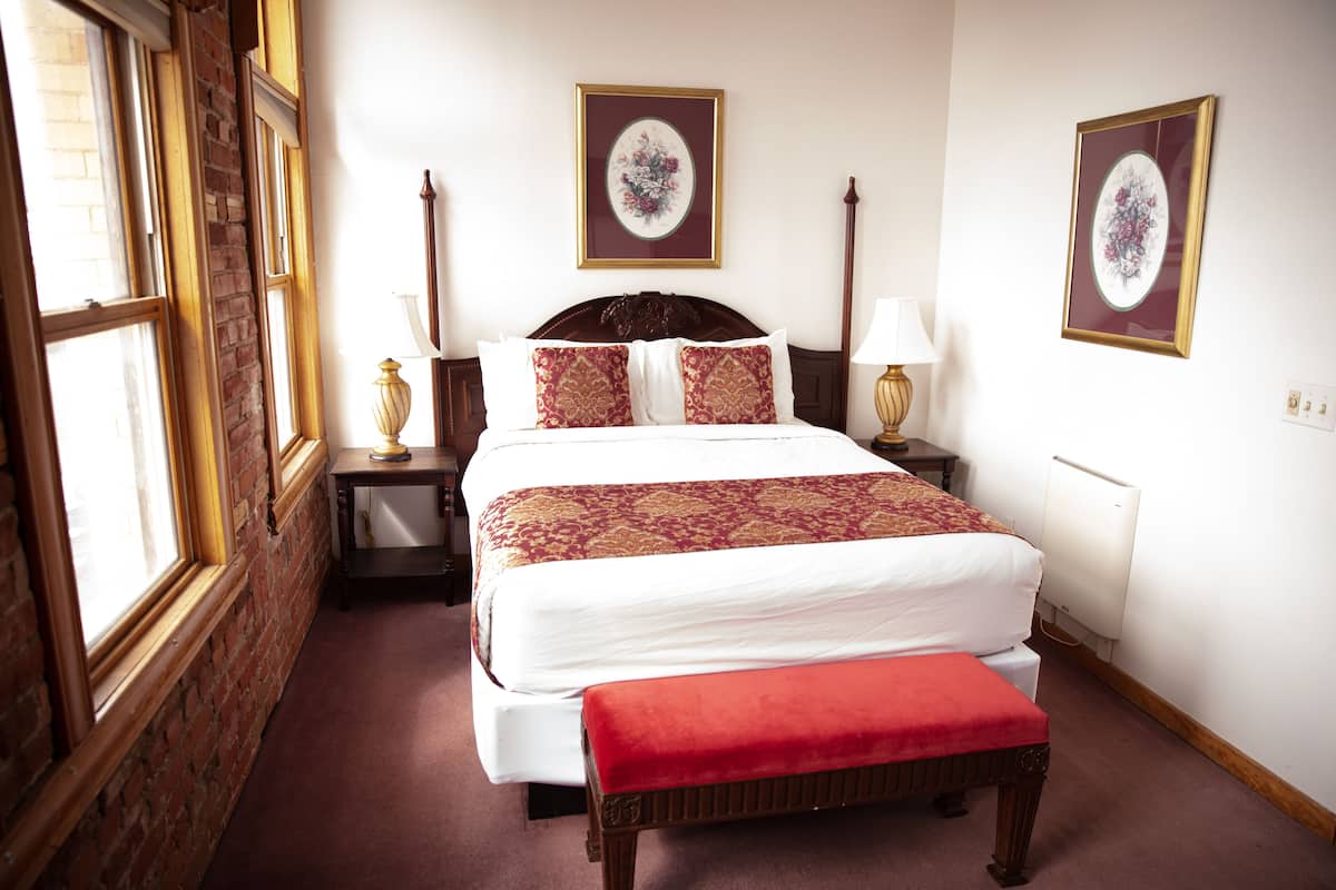 A neatly arranged hotel room with a large bed, a bedside table, a nightstand, a red bench, and two framed floral artworks on the wall.