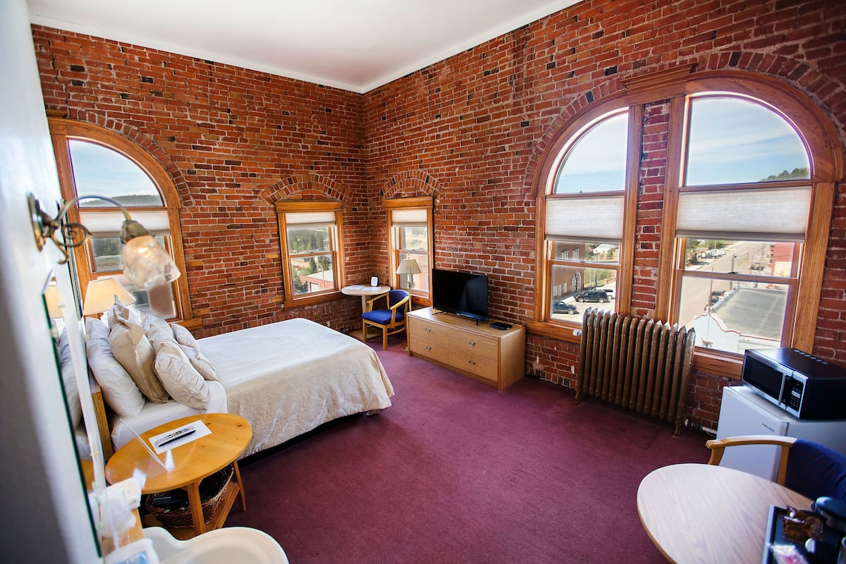 A room with a brick wall, a bed, a TV, a desk, and a view of the city.