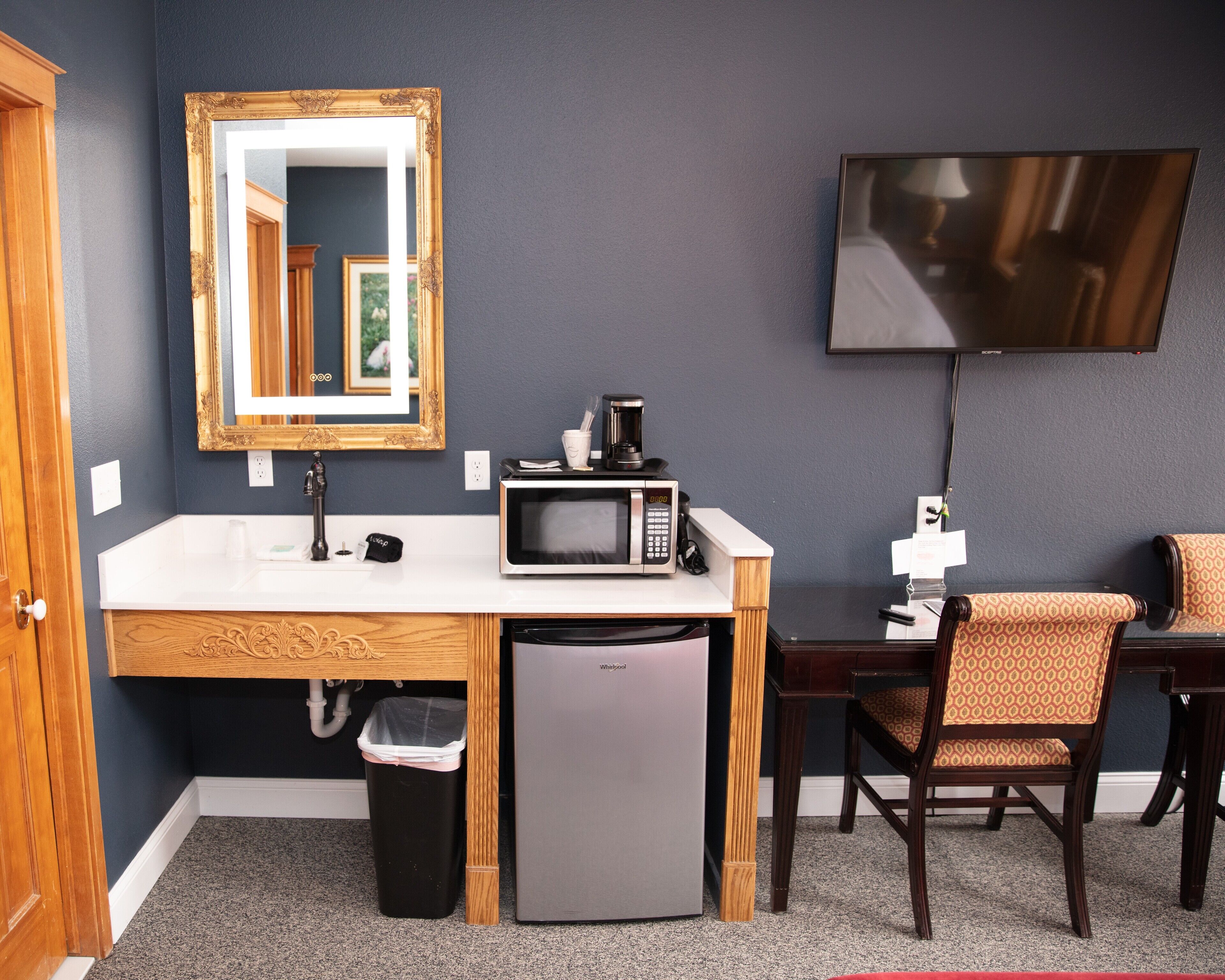 A compact hotel room with a kitchenette, a microwave, a small refrigerator, a sink, a mirror, a television, and two patterned chairs.
