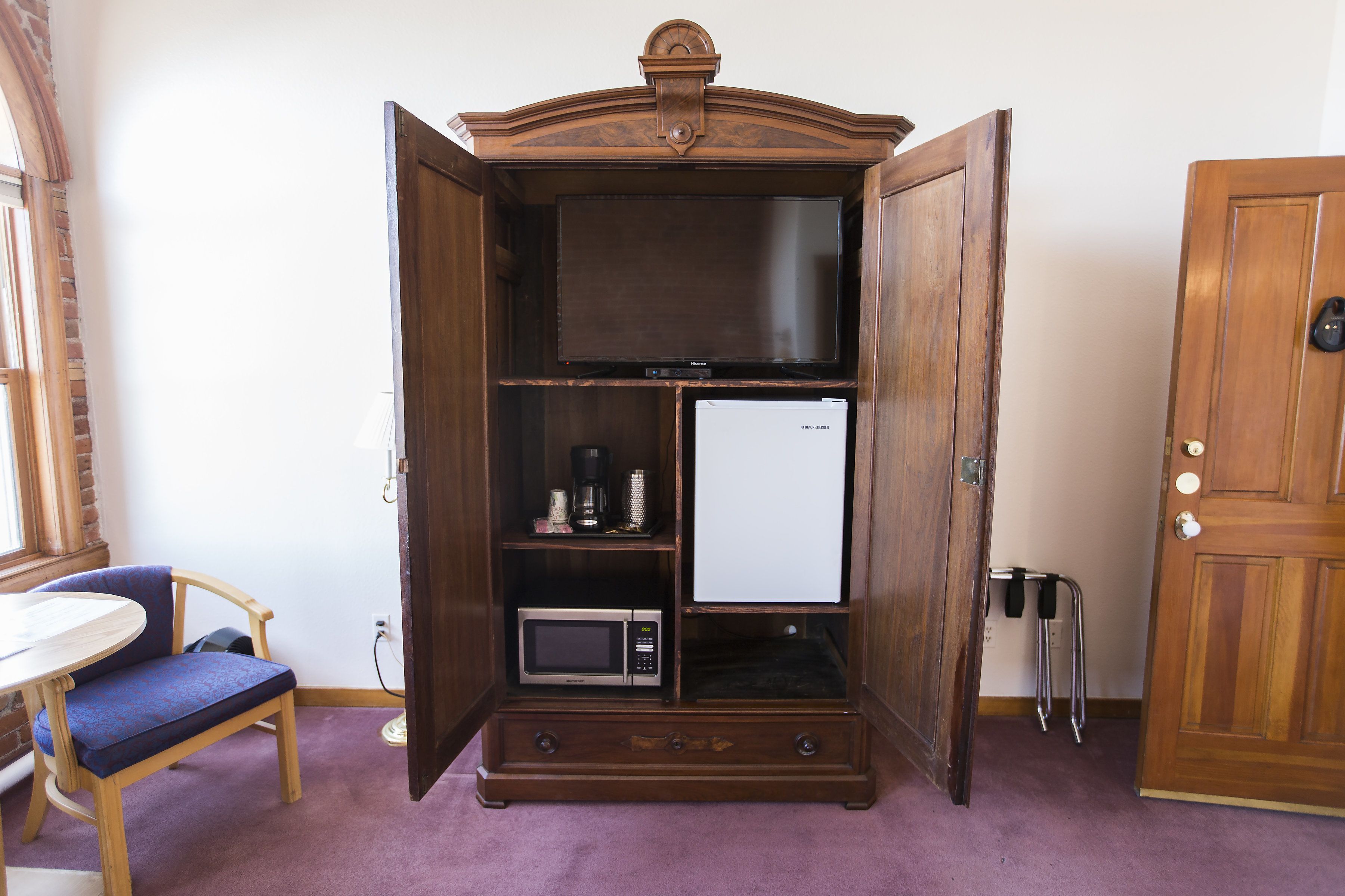 Habitación compacta con un mueble de madera que contiene un microondas, un refrigerador pequeño y una cafetera. A la izquierda, hay una silla y una mesa, y a la derecha, una puerta.