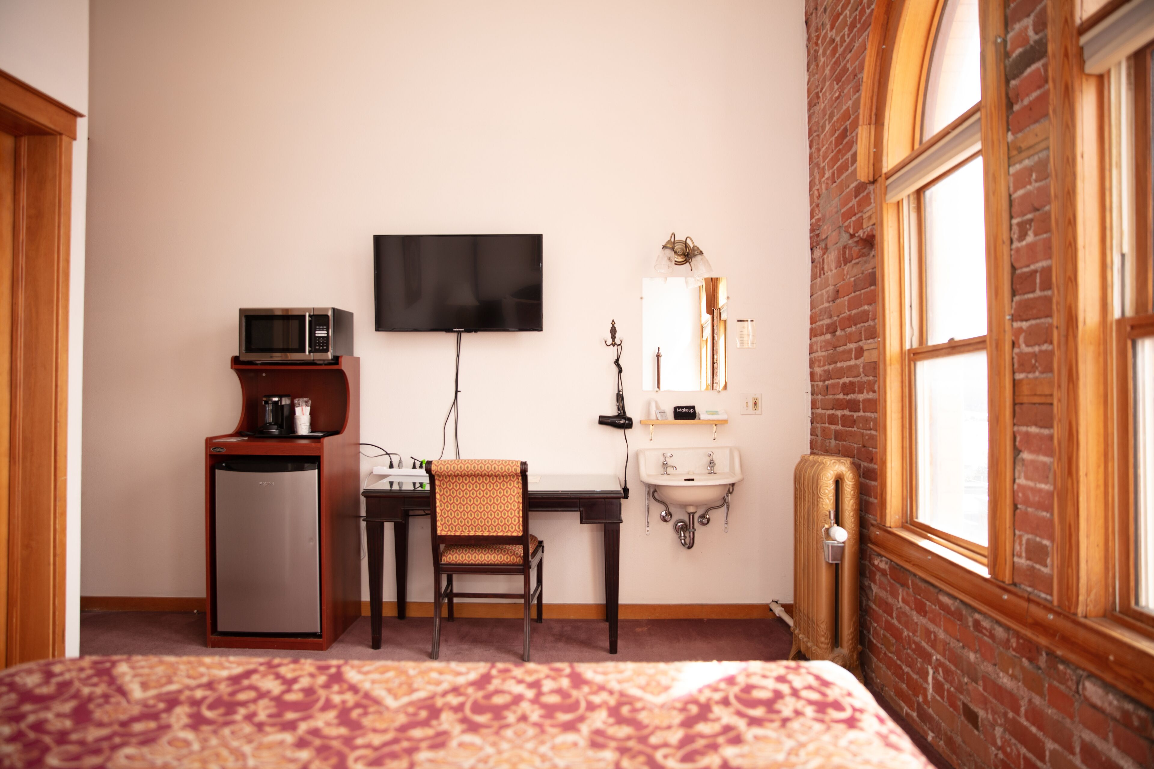 Une chambre d’hôtel comprenant un lit, un bureau avec une chaise, un four à micro-ondes, une télévision, un lavabo et une fenêtre donnant sur un mur en briques.