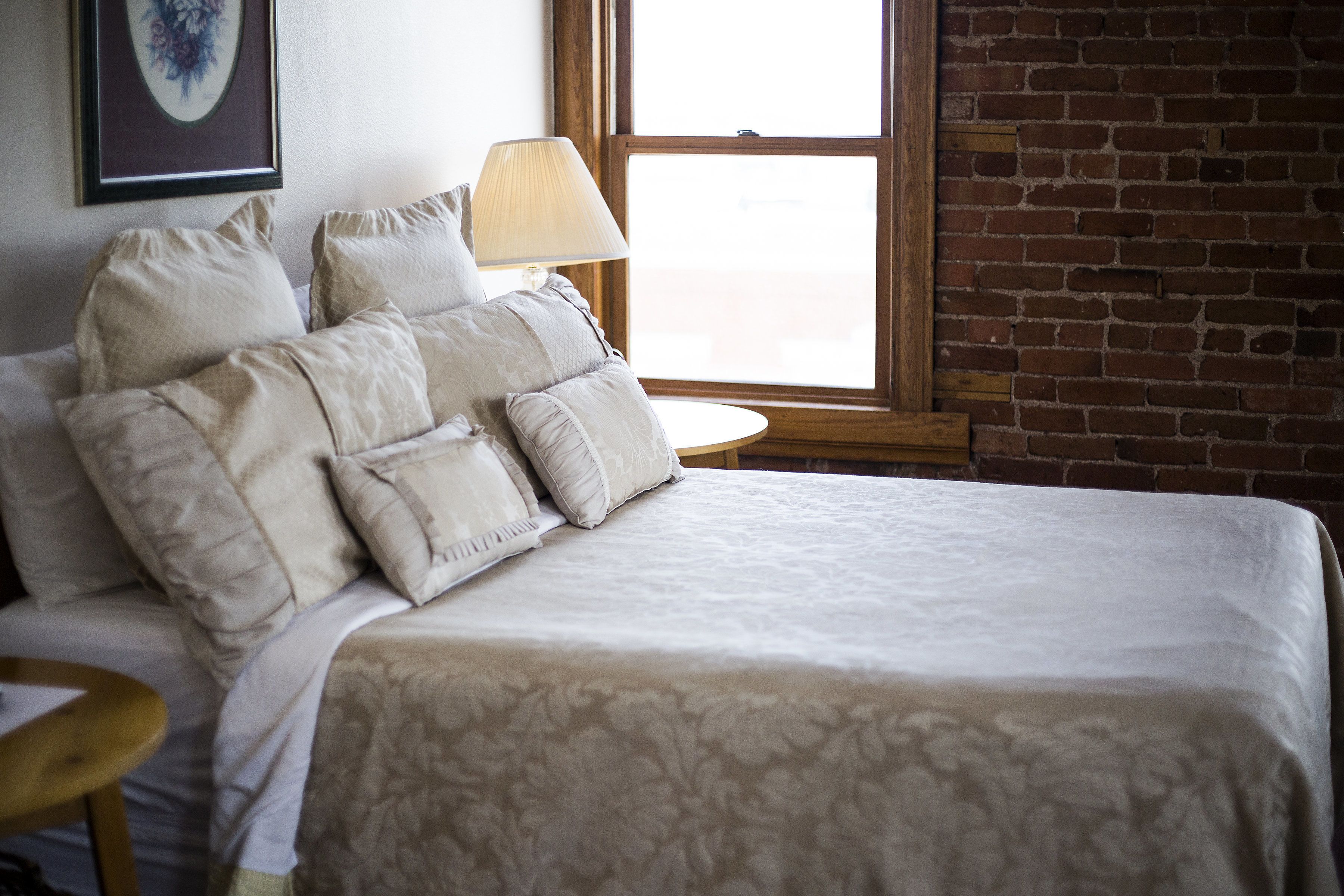 Una cama bien hecha con una colcha estampada, una lámpara de noche y vista a un muro de ladrillo a través de una ventana.