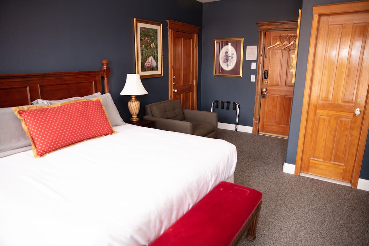 A hotel room with a bed, a chair, a bench, a lamp, and two doors.