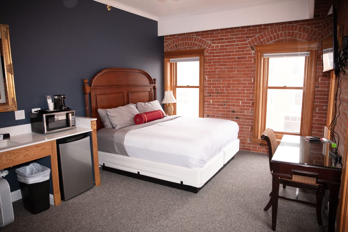 A hotel room with a bed, a desk with a microwave, a trash bin, and a brick wall.
