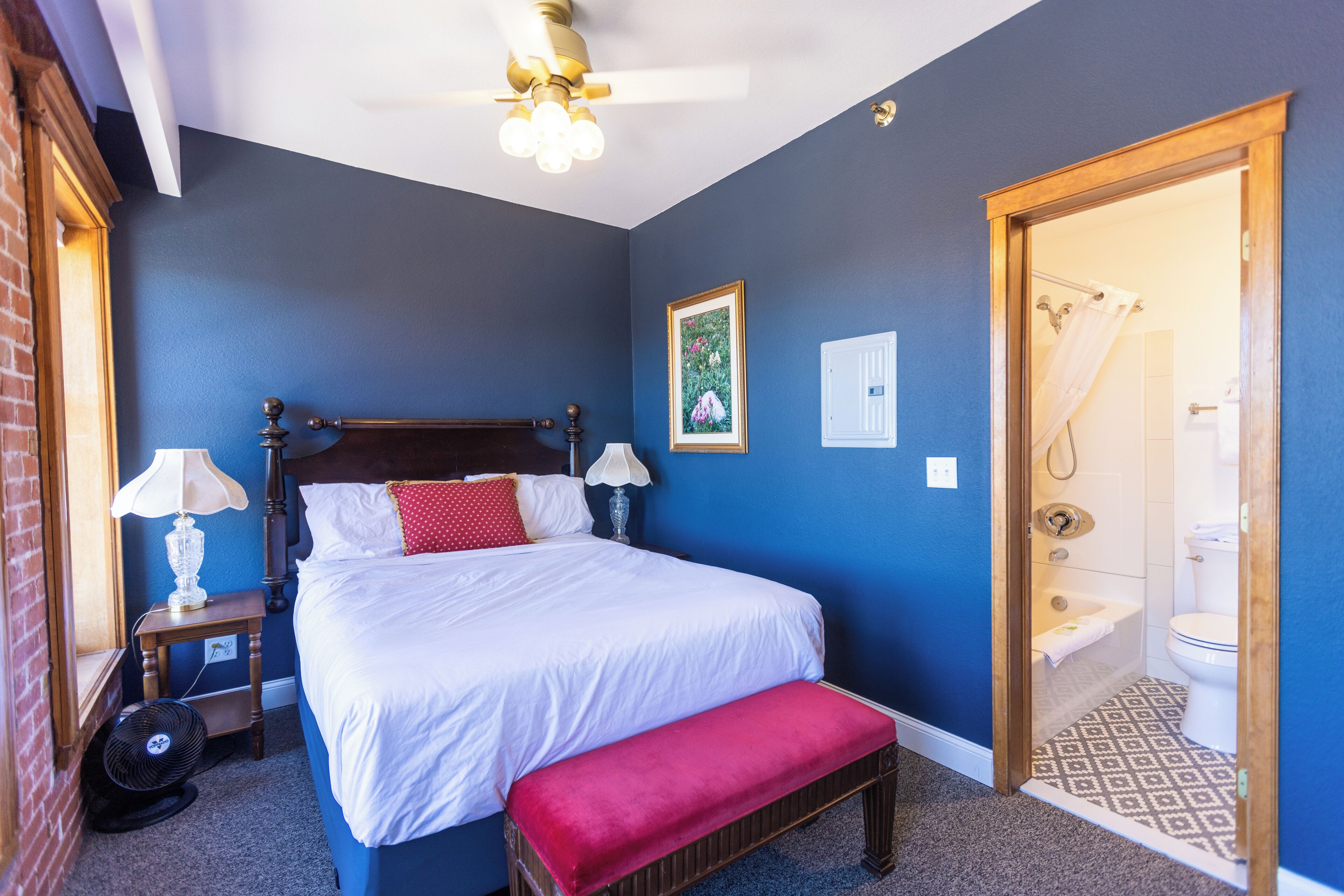 A bedroom with a bed, bedside lamps, a fan, a bench, a ceiling fan, a framed picture, and a bathroom visible through an open door.