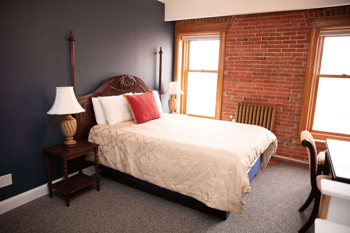 A bedroom with a bed, two bedside tables, a chair, a window with a view of a brick wall, and a radiator.