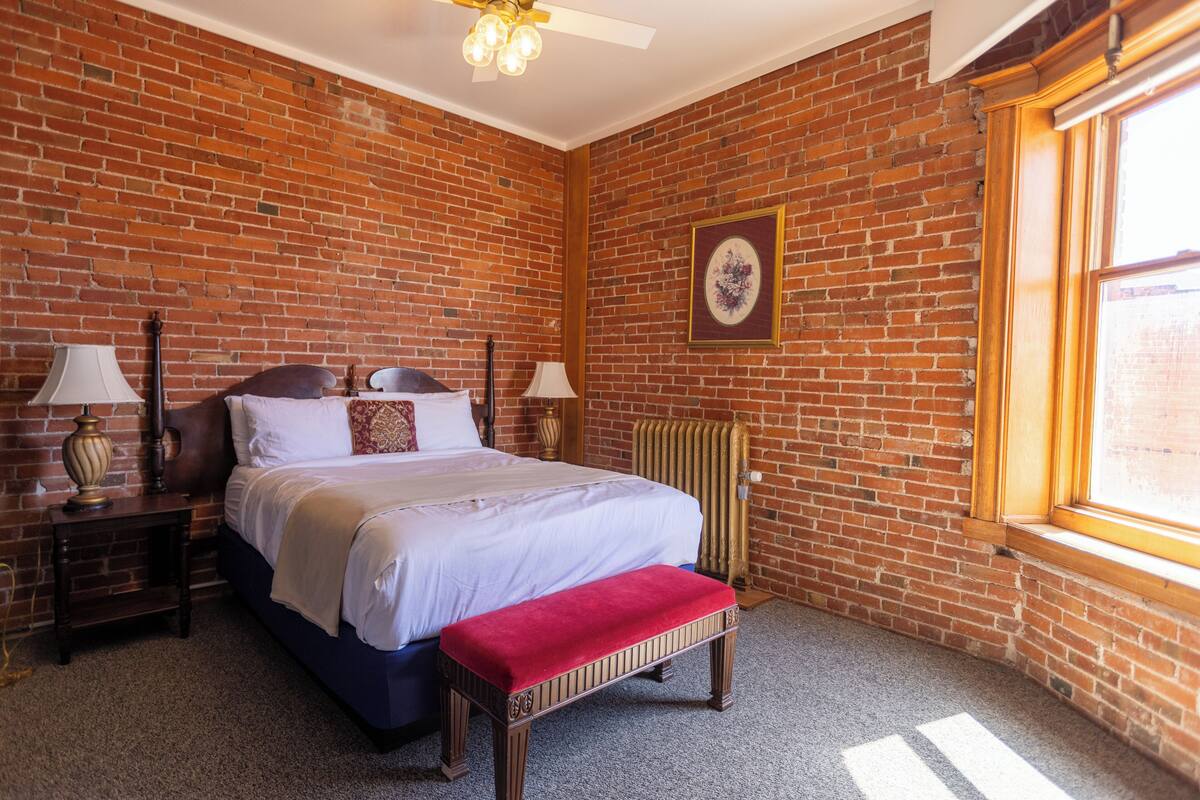 A room with a brick wall, a bed with a red bench, two bedside tables with lamps, and a radiator.