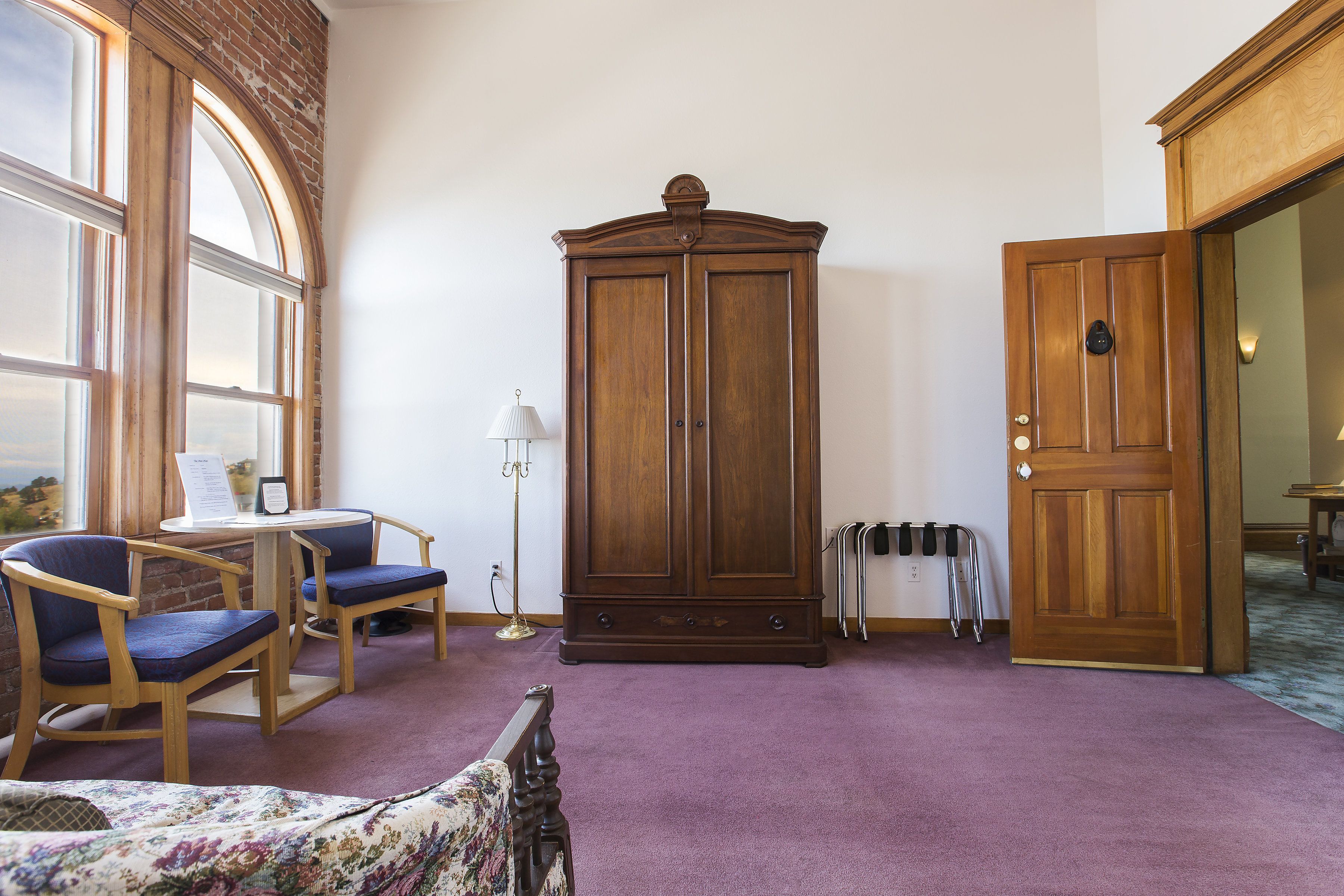 Une pièce comprenant une grande armoire en bois, deux chaises, une petite table et un lit recouvert d’une couvre-lit à motifs floraux.