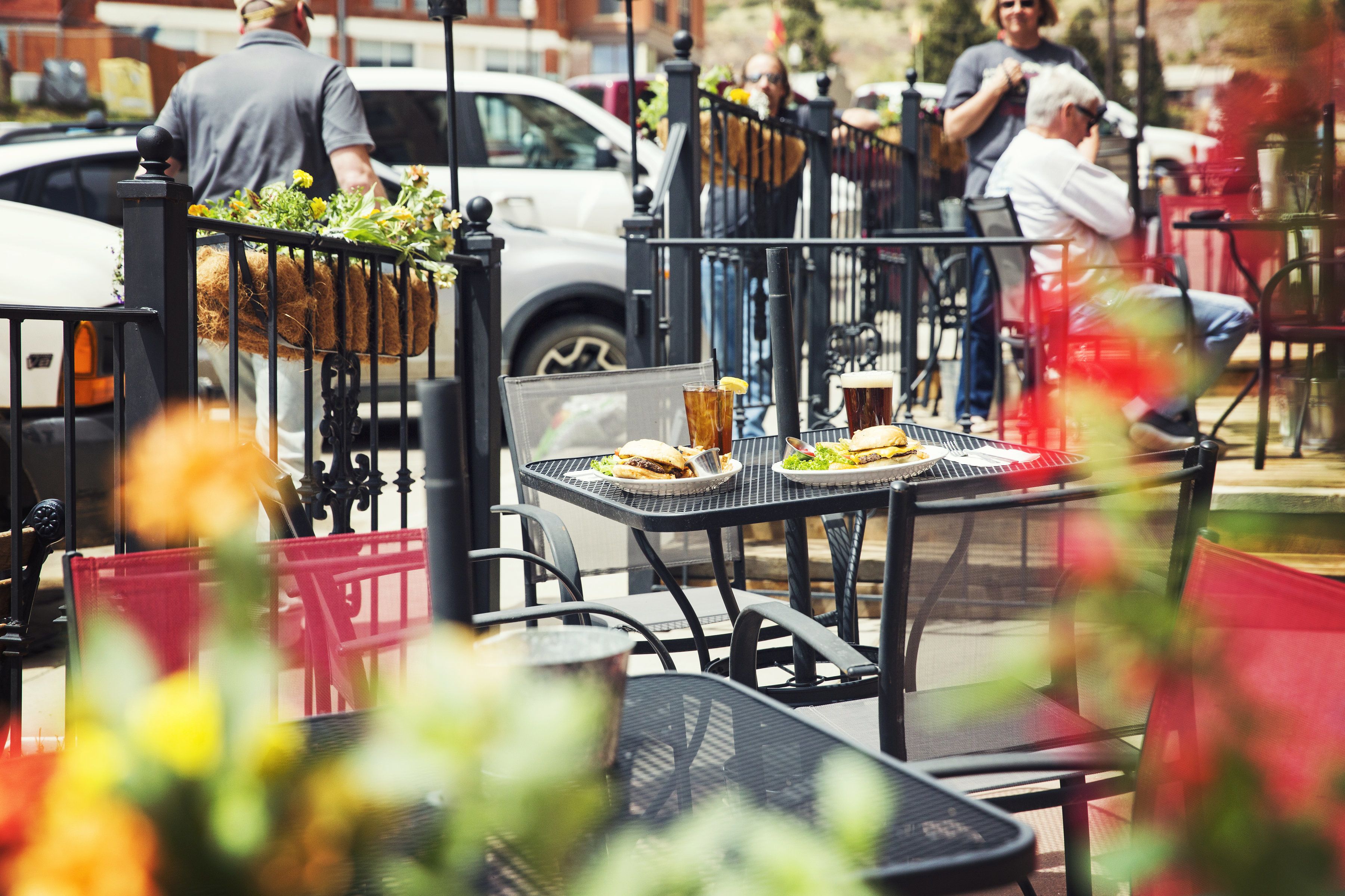 An outdoor dining area with tables and chairs, a car parked nearby, and a fence with greenery.