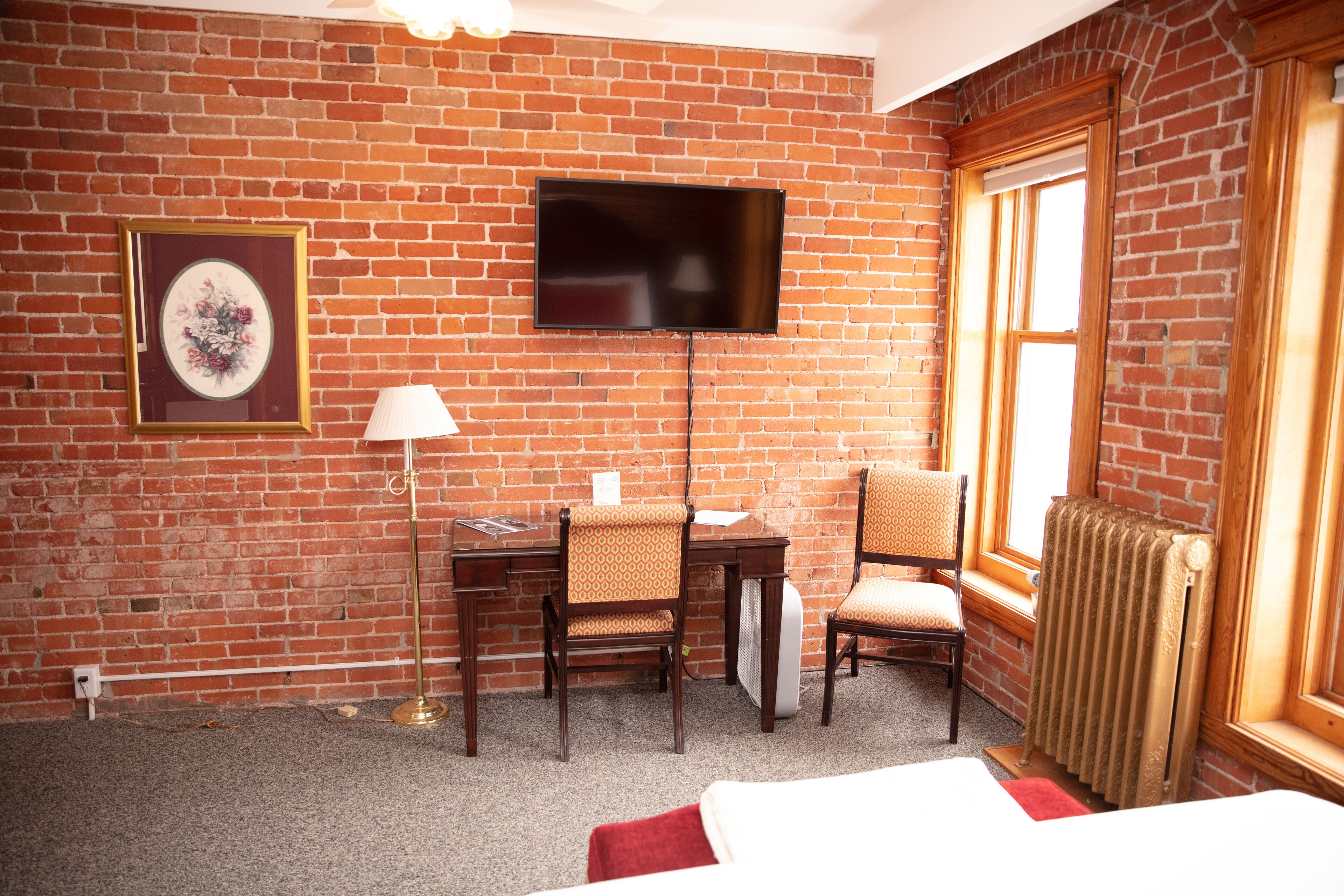 Ett hotellrum med en tegelvägg, en platt-TV som hänger ovanför ett skrivbord i trä, två mönstrade stolar, ett element och en inramad tavla på väggen.