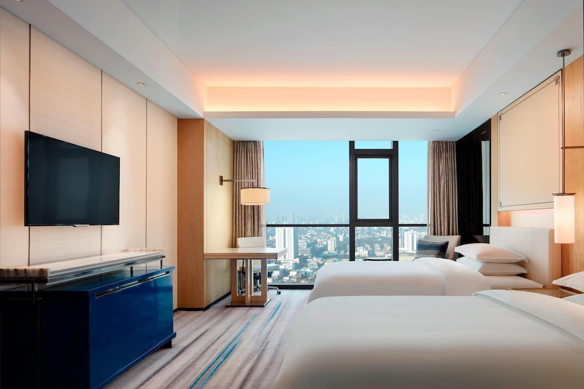 Ein Hotelzimmer mit einem großen Bett, einem Fernseher, einem Schreibtisch und Blick auf die Stadt.
