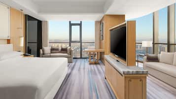 A hotel room with a large bed, a sofa, a TV, and a desk. There is a large window with a city view.