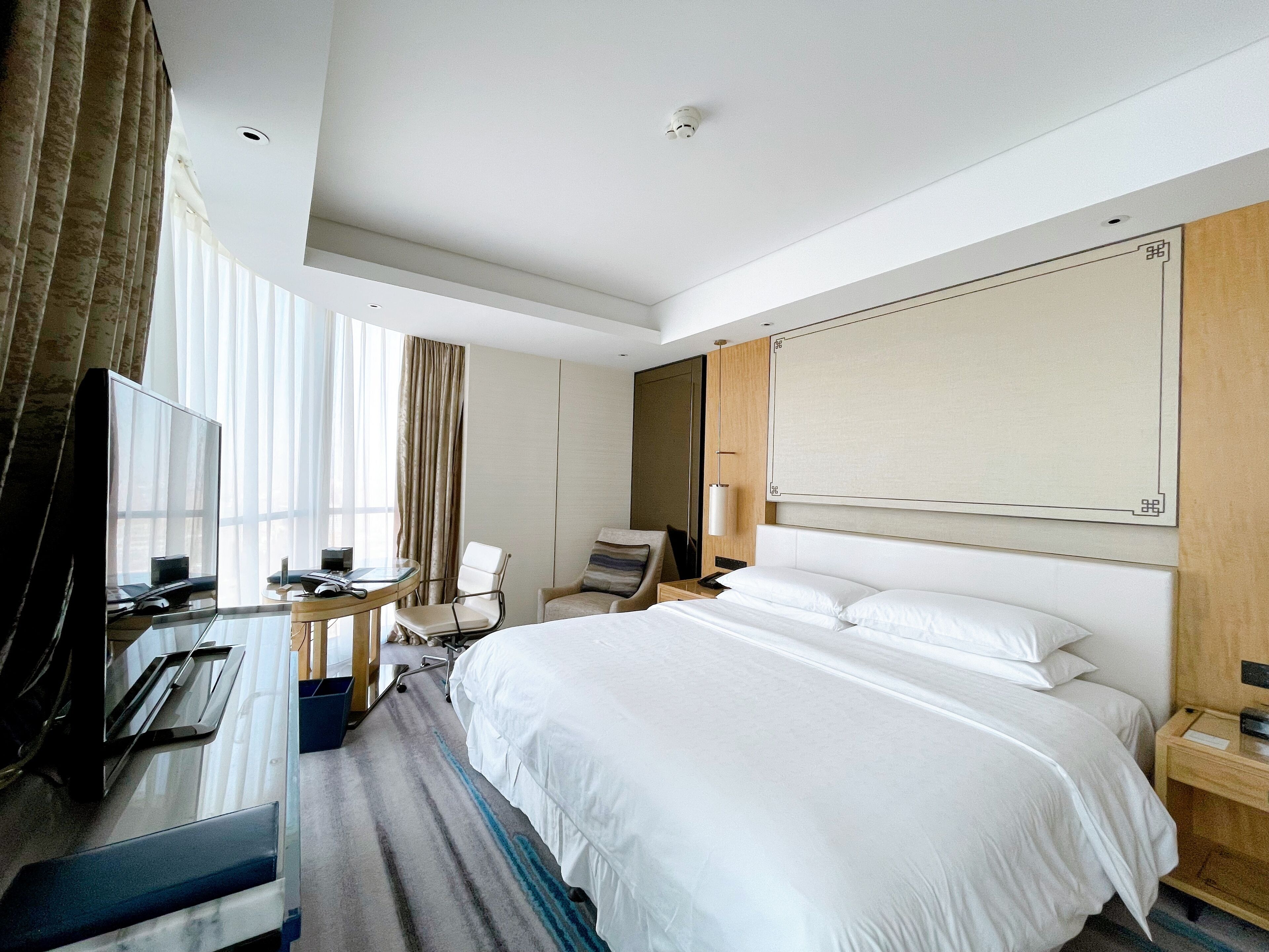 A hotel room with a bed, a desk with a computer, a chair, a TV, and a large window with curtains.