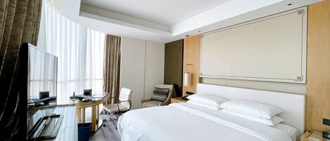 A hotel room with a bed, a desk with a computer, a chair, a TV, and a large window with curtains.