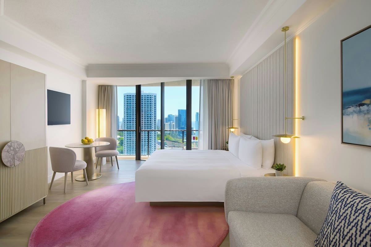 Een hotelkamer met een bed, een bank, een bureau en een stoel. Grote ramen bieden uitzicht op de stad.