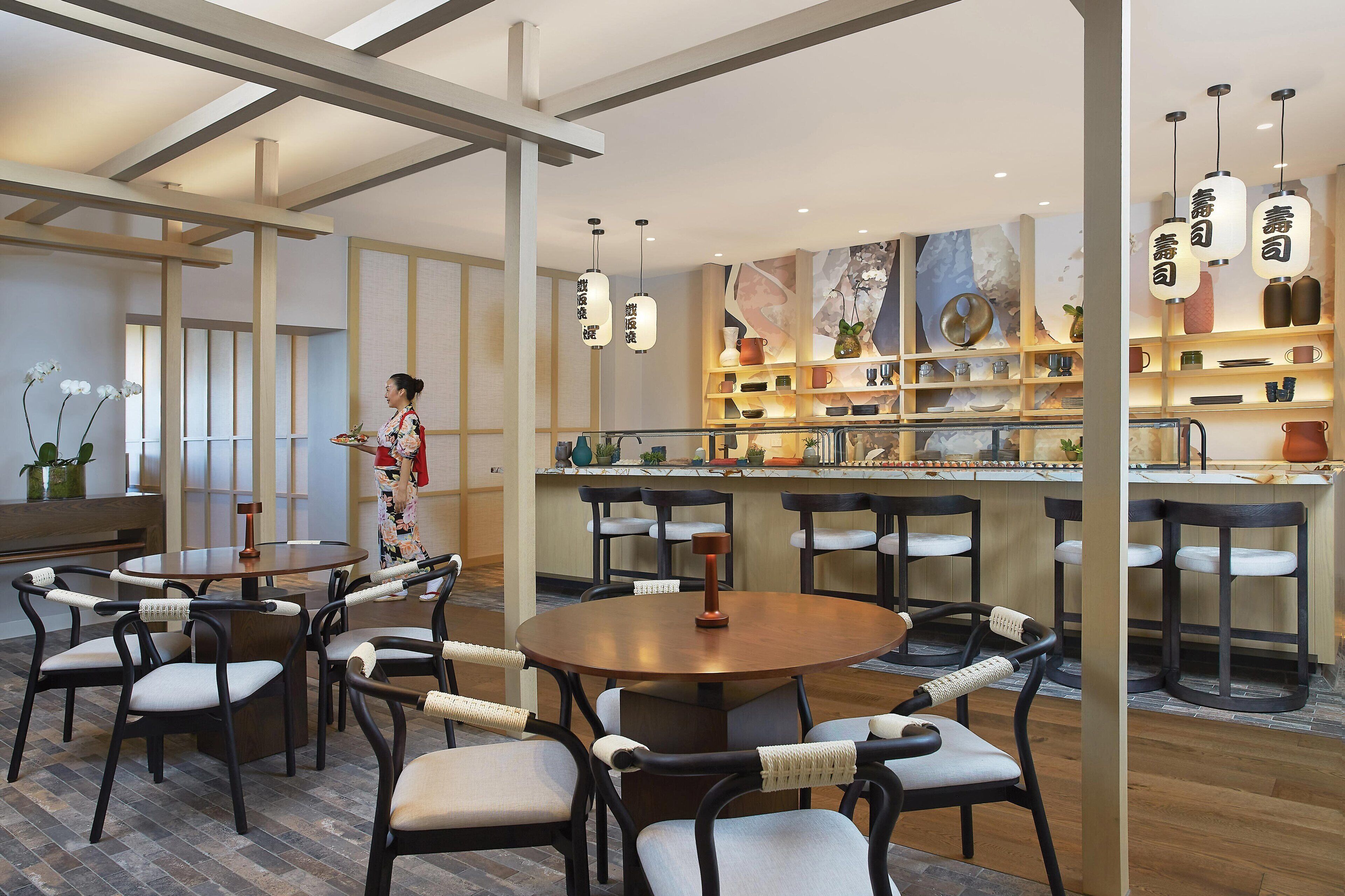 Een modern restaurantinterieur met ronde tafels en stoelen, een bar met schappen vol servies, en hangende pendantlampen.
